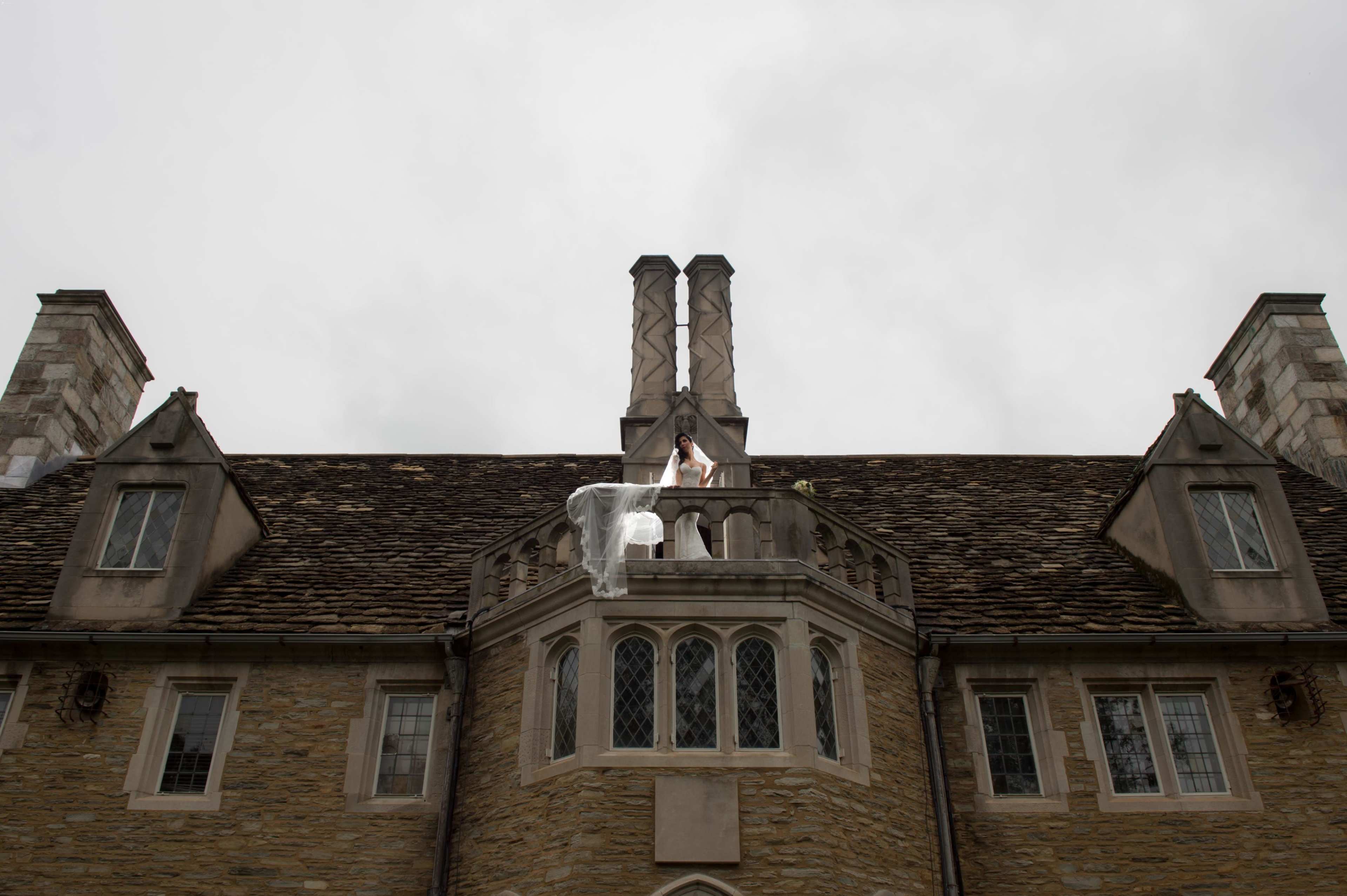 A woman in a white dress stands on the balcony of a stone mansion with two prominent chimneys.