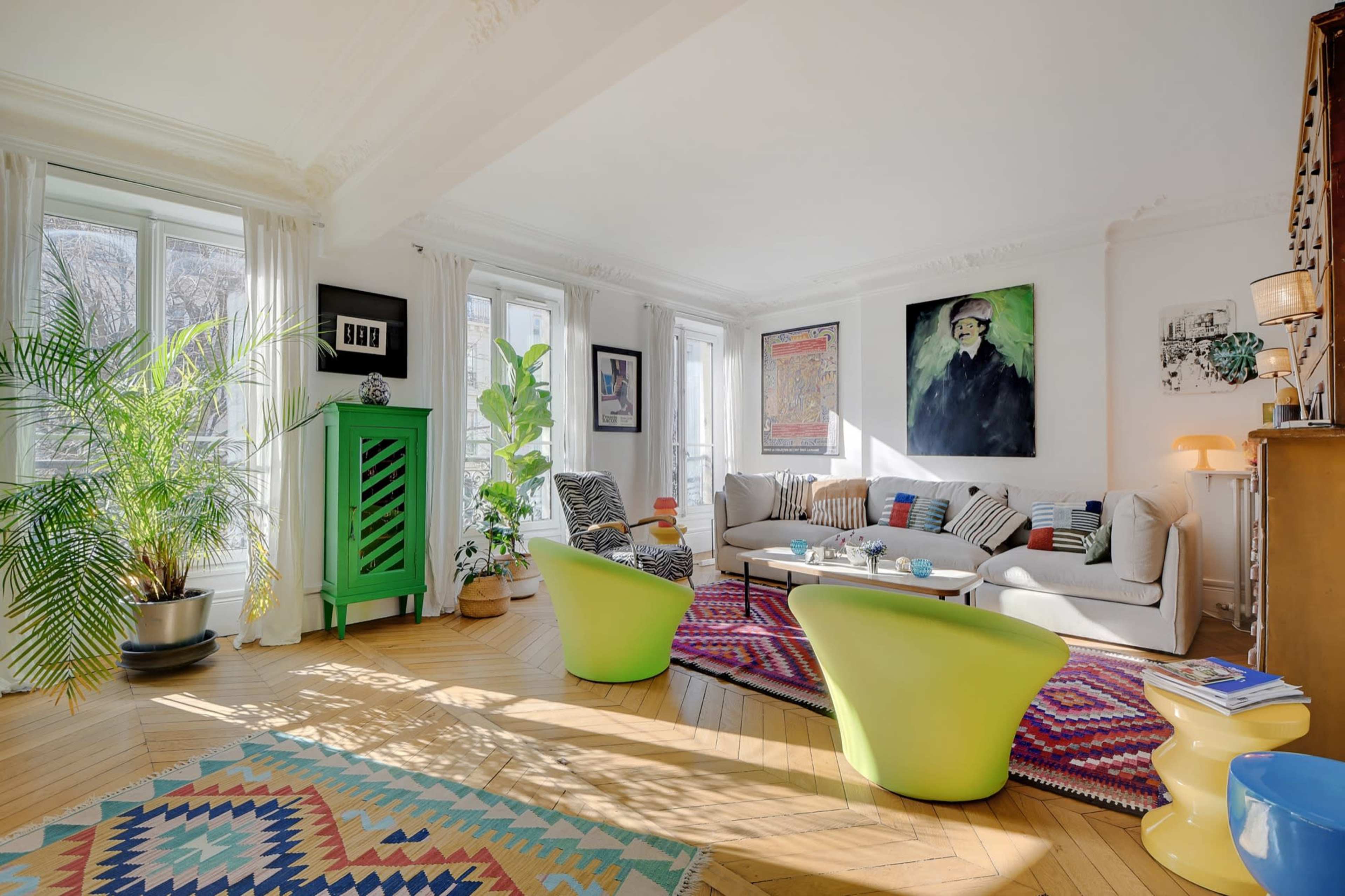 A brightly lit living room featuring a green cabinet, a colorful rug, a sofa with various cushions, and large windows with plants.