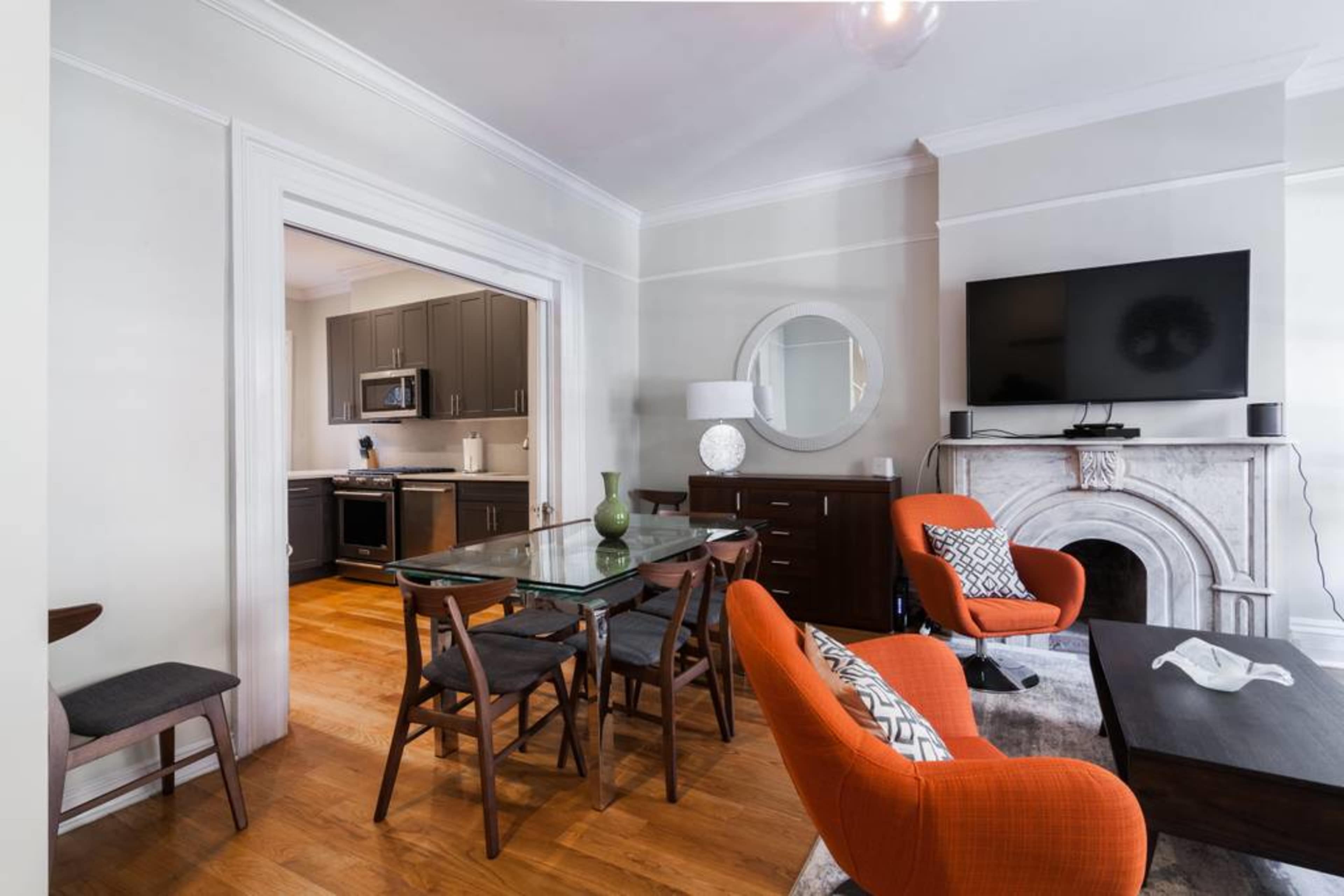 A living room with a dining area features an orange armchair, a glass dining table, and a modern kitchen visible in the background.
