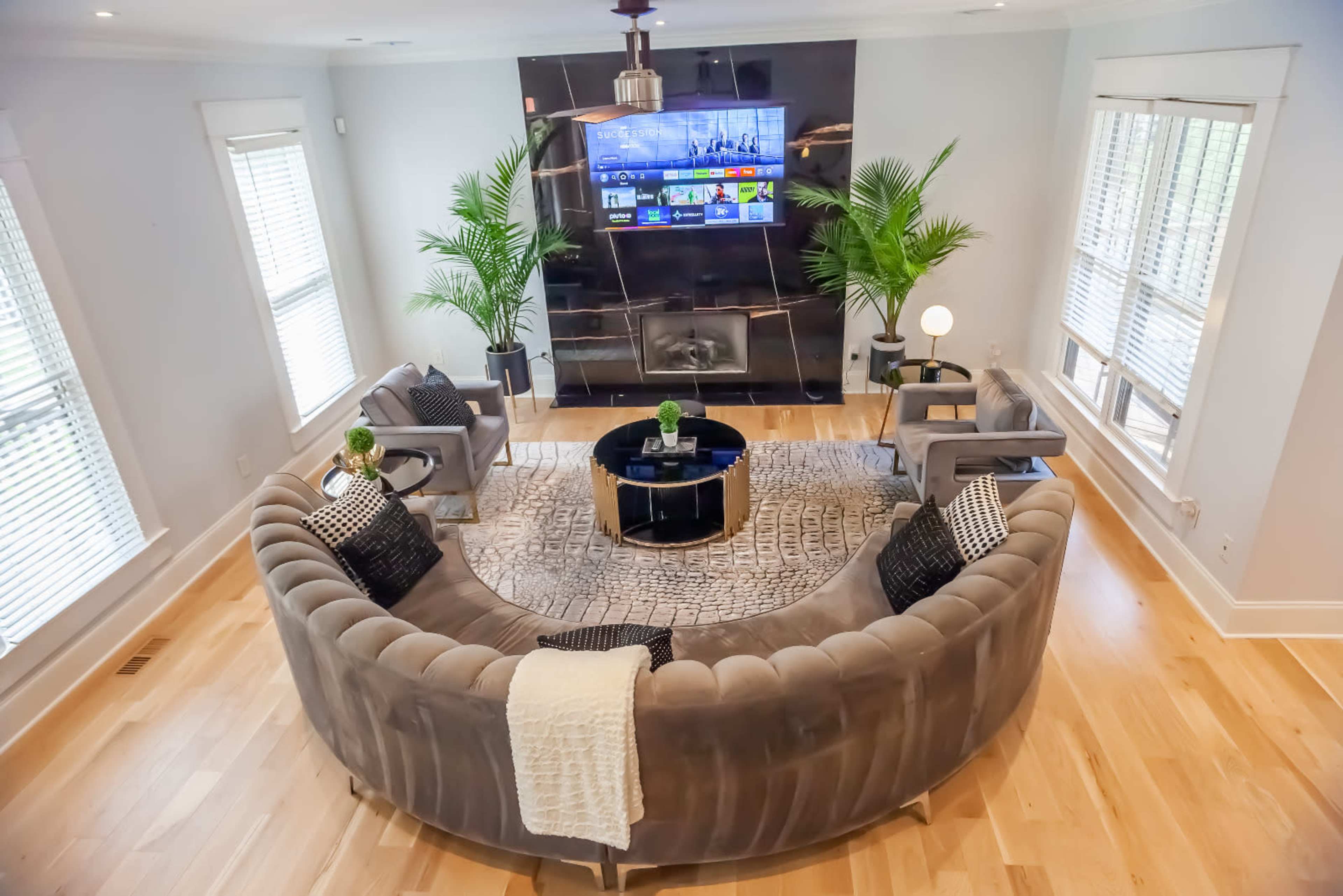 A modern living room features a curved grey sofa, a round coffee table, and a wall-mounted television, surrounded by potted plants and large windows.