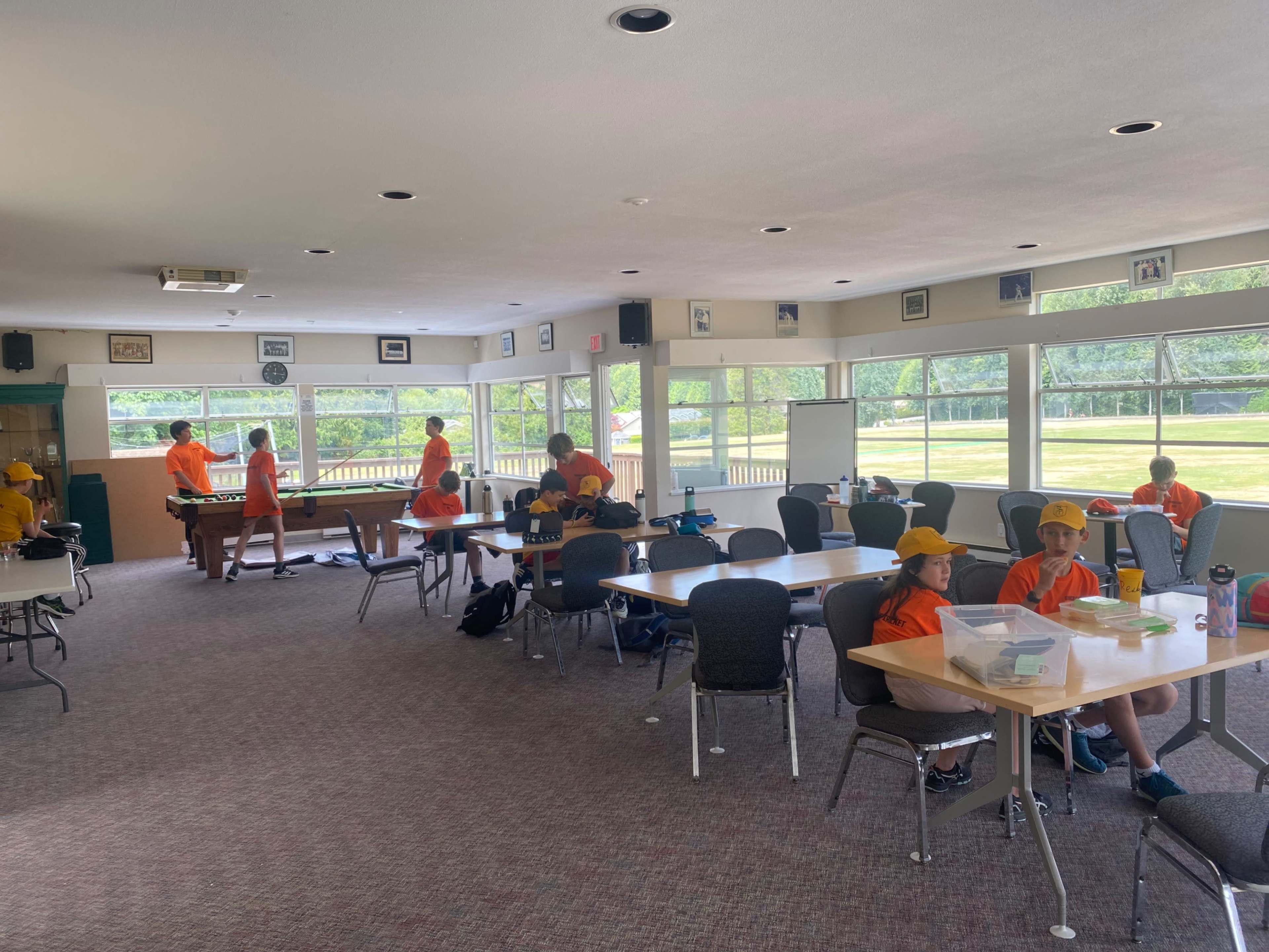 A group of children in orange shirts and yellow caps are engaged in various activities inside a bright room with large windows and several tables.