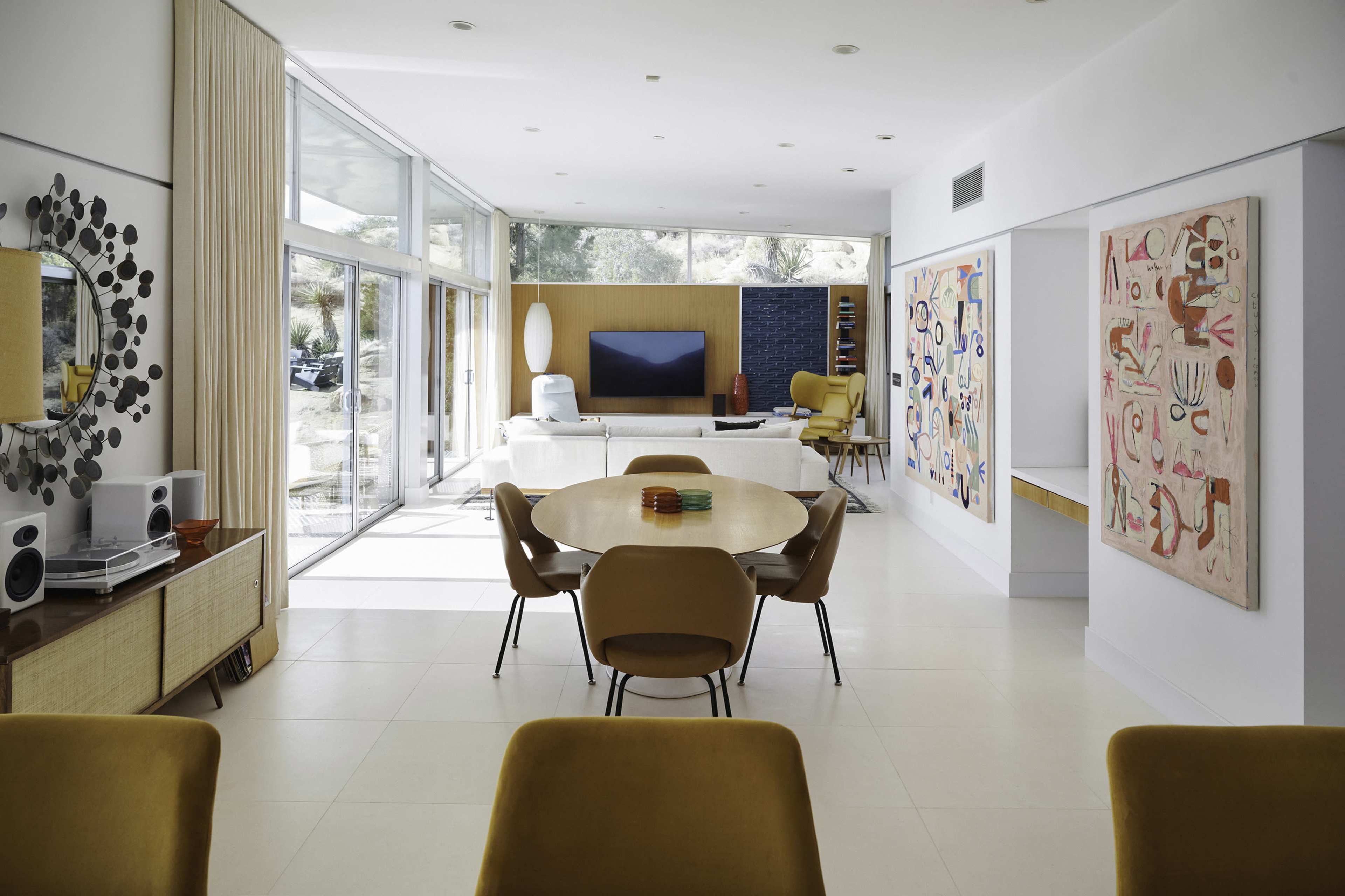 A modern living space featuring large windows, a round dining table surrounded by chairs, and colorful artwork on the walls.