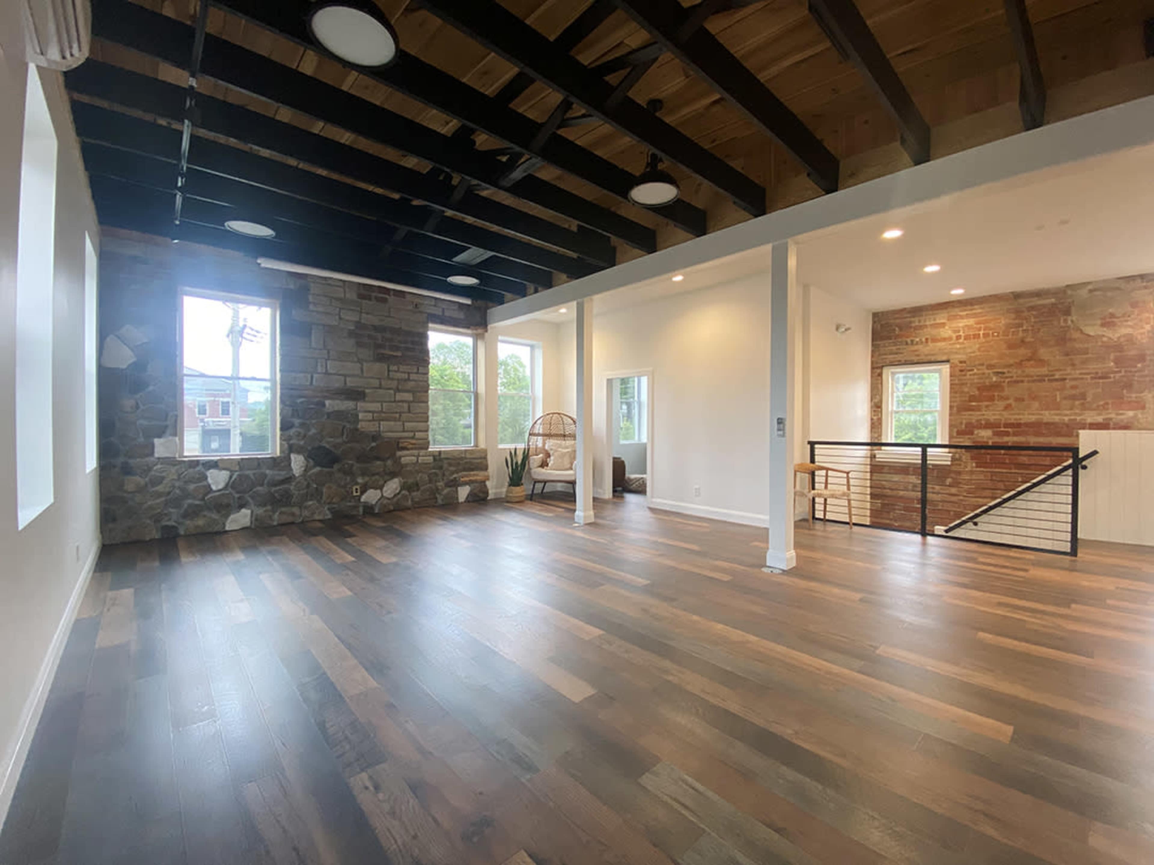 A spacious room with wooden floors, exposed beams, a stone wall, and large windows allowing natural light to enter.