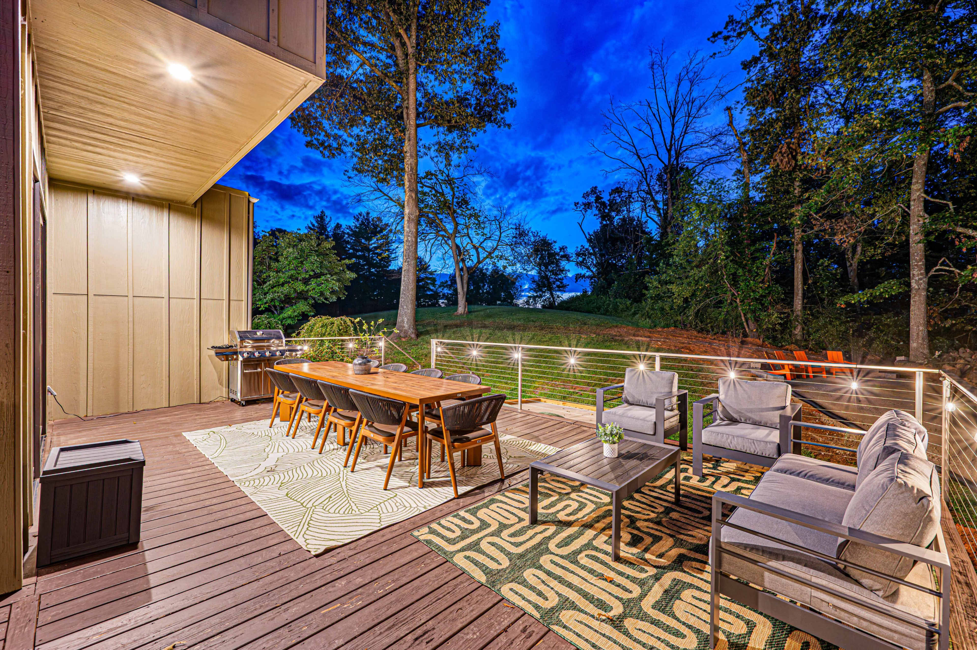 A spacious deck featuring a dining table, seating area, and a grill, surrounded by trees under a twilight sky.