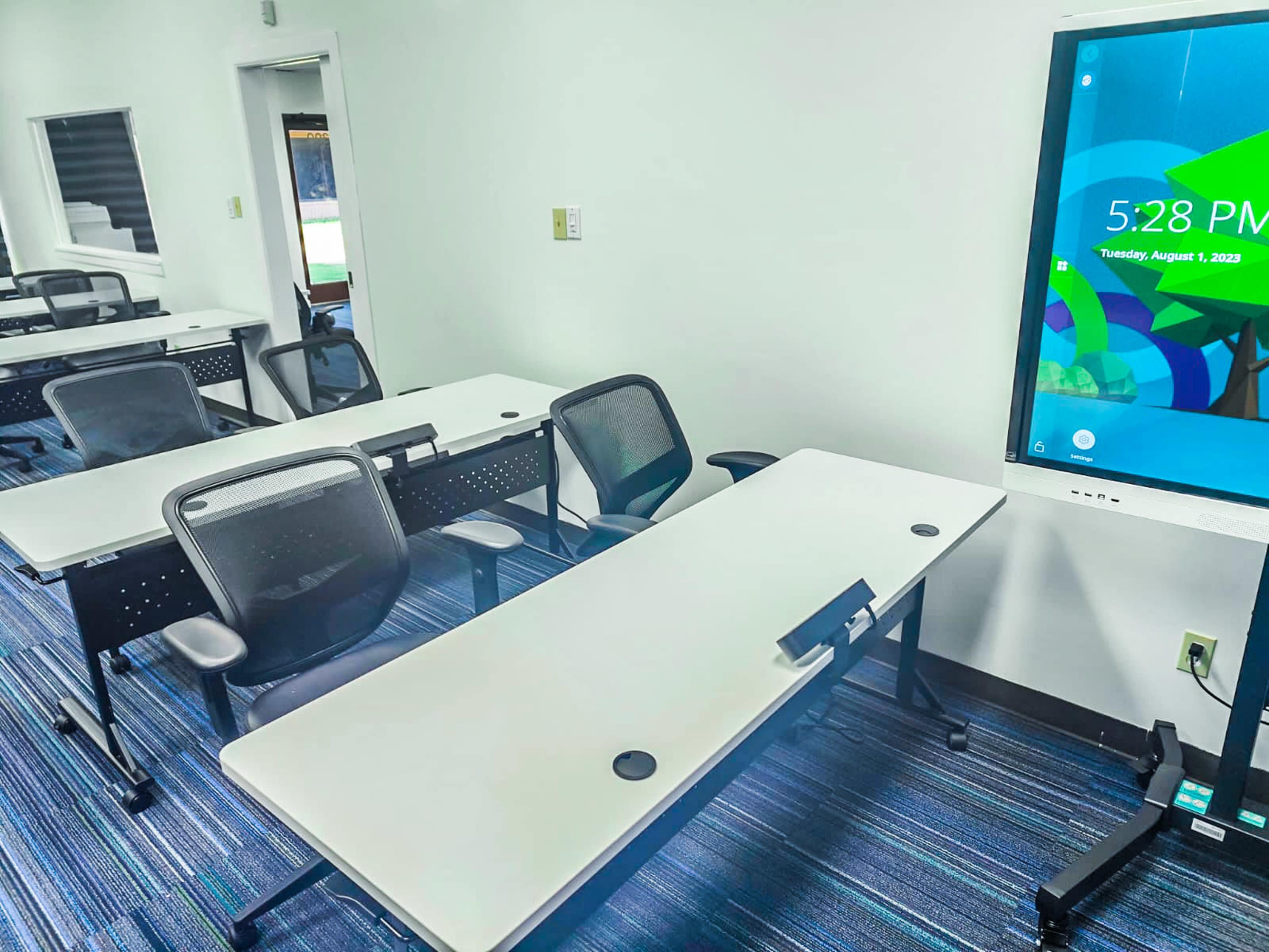 A classroom is set up with several white tables and black chairs, alongside a digital display showing the time and date.