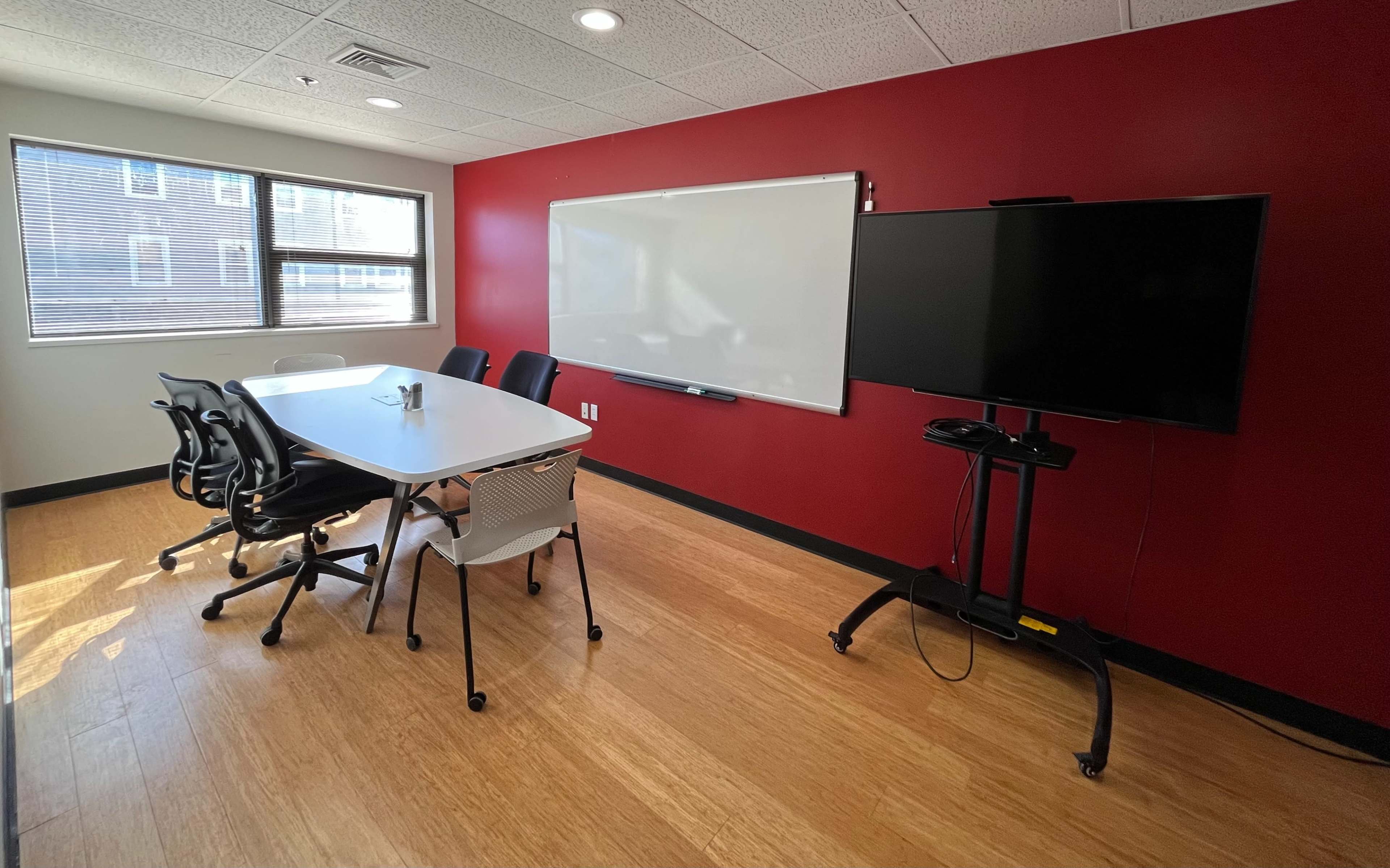 The image shows a small conference room with a whiteboard, a television on a stand, and a table surrounded by chairs.