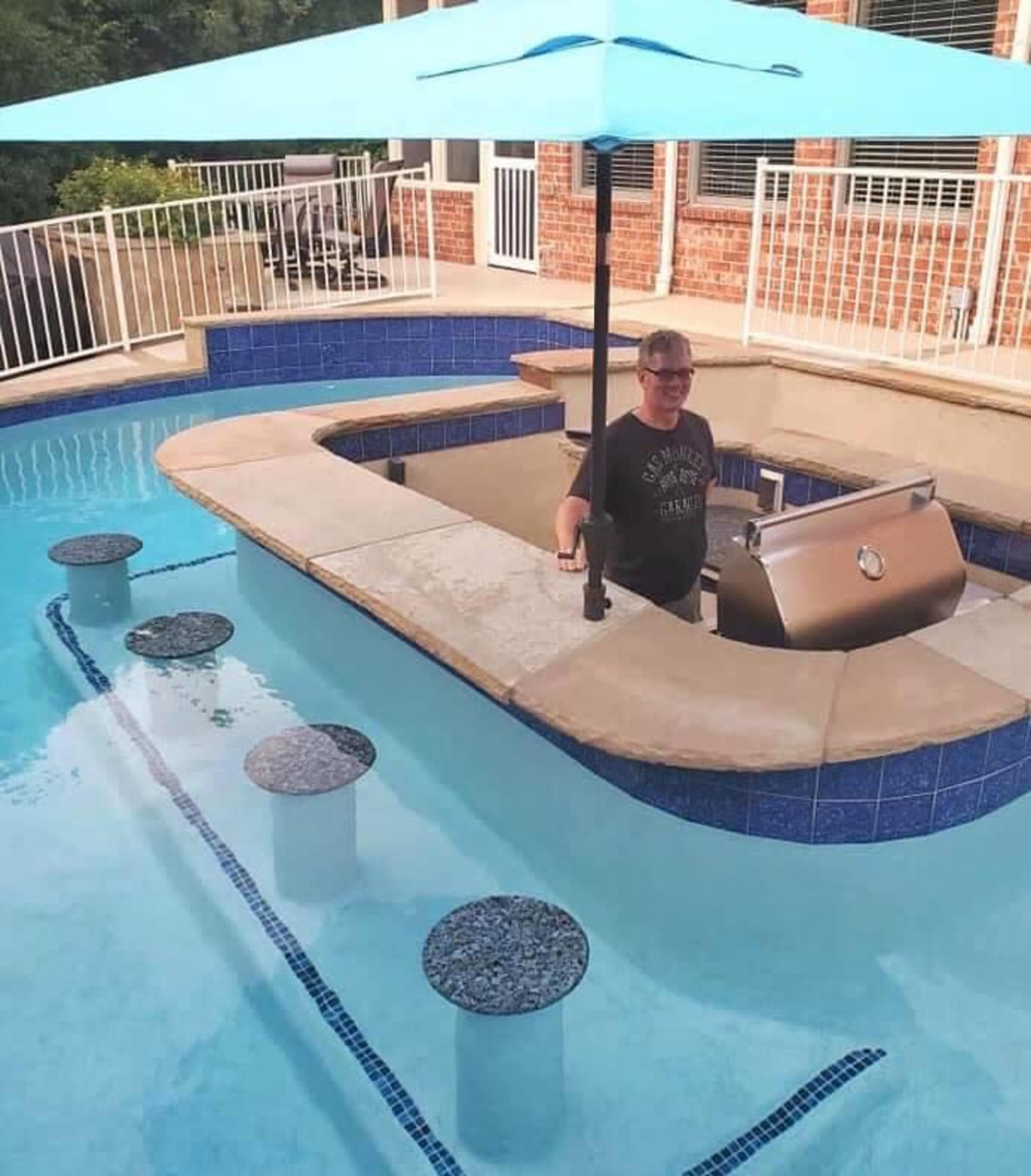 A person stands at a bar-style grill in a swimming pool, shaded by a blue umbrella.