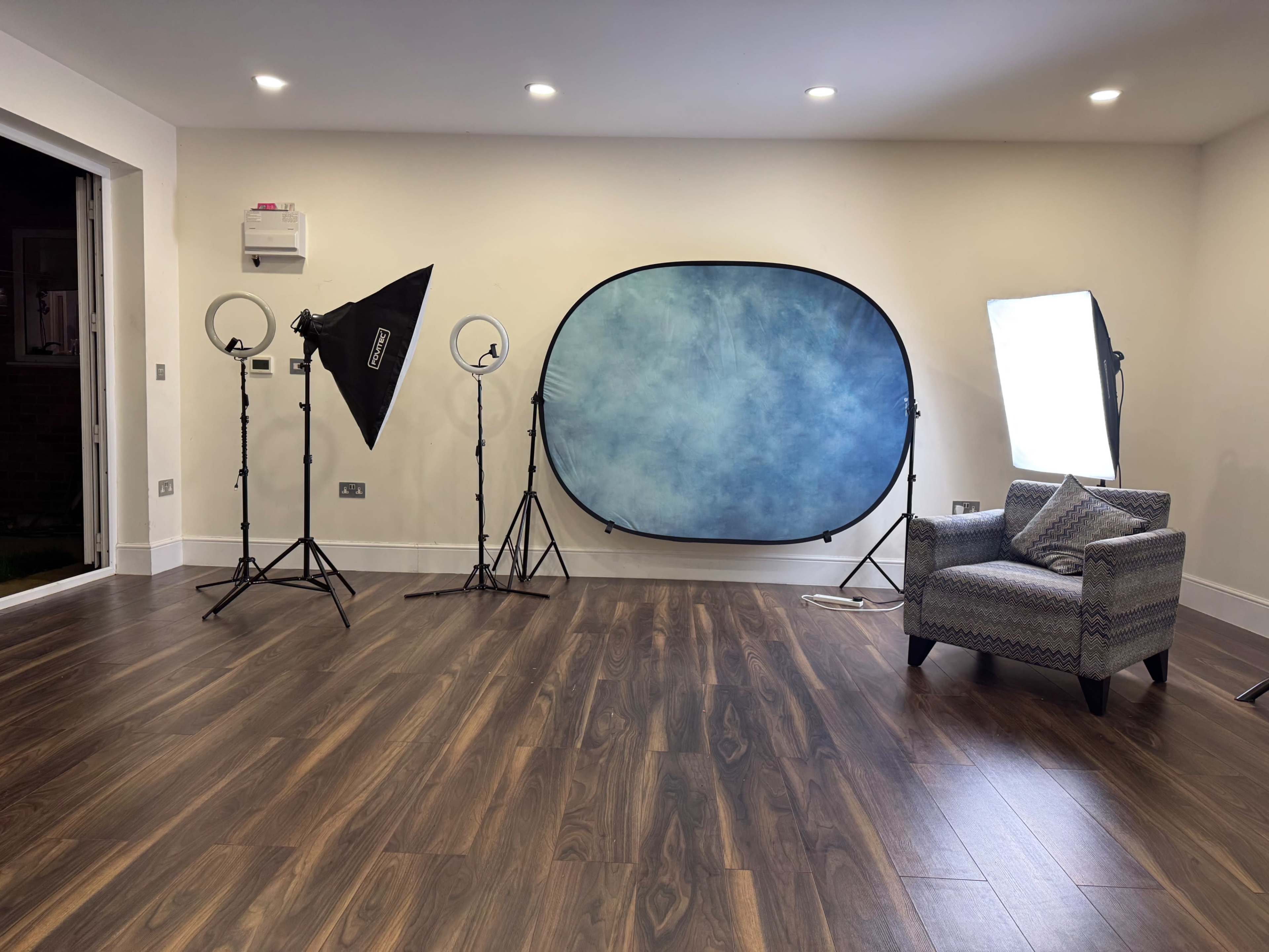 The image shows a photography studio setup featuring a backdrop, lighting equipment, and a chair on a wooden floor.