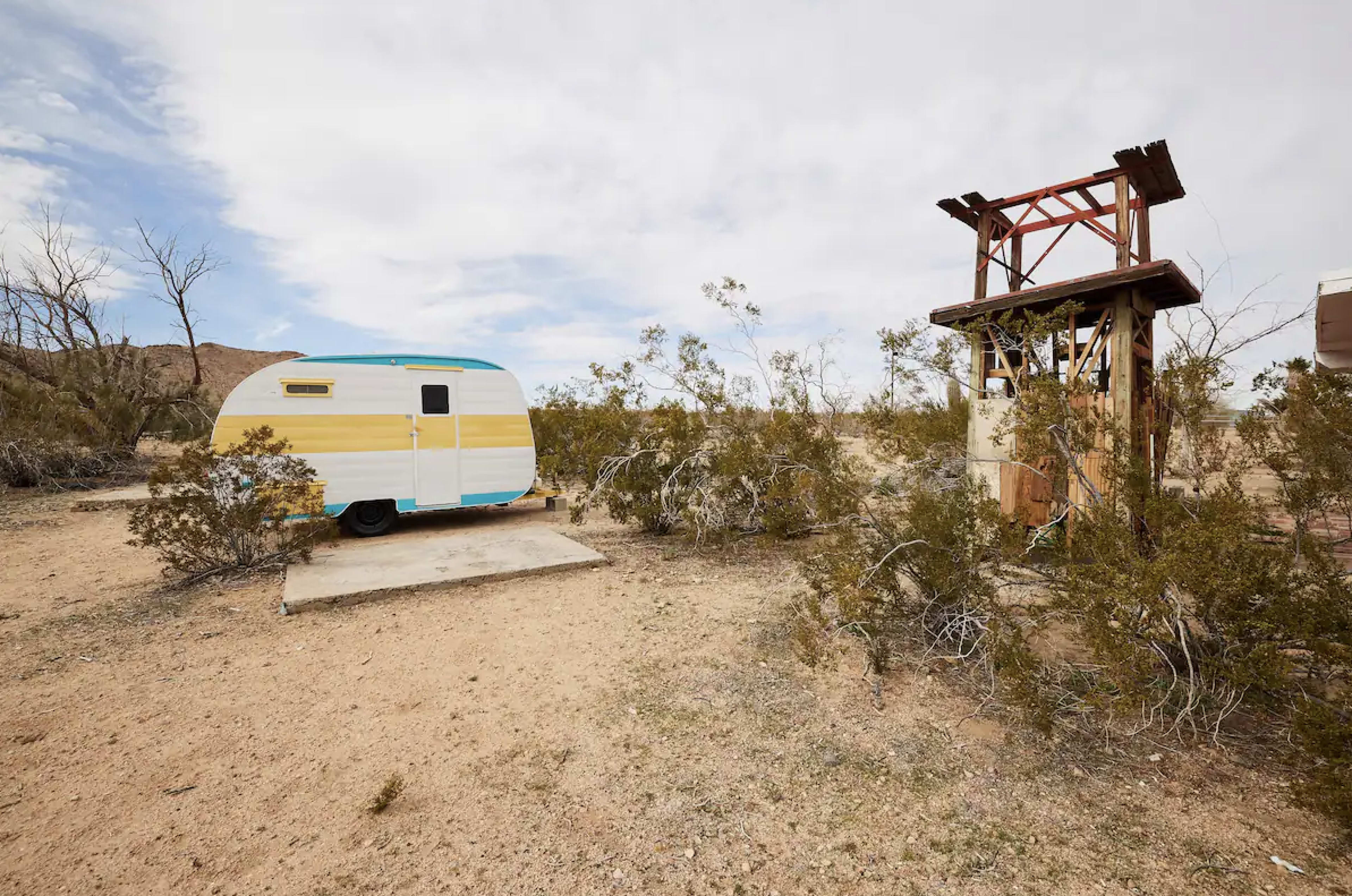 A colorful vintage caravan is parked on a concrete slab next to a wooden structure in a desolate landscape with sparse vegetation.