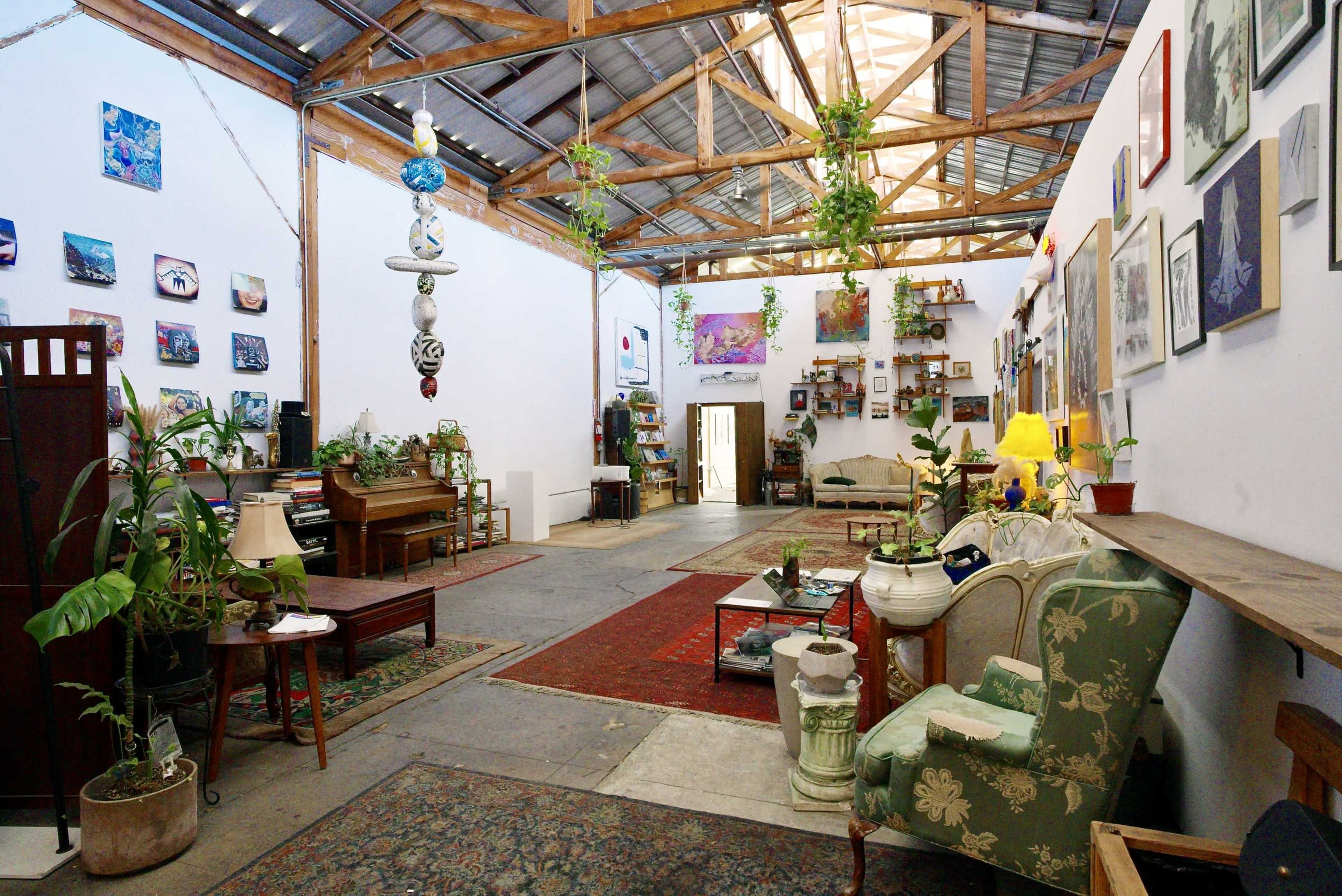 The image shows a spacious, open interior with wooden beams, various plants, artwork on the walls, and a mix of furniture including a piano, couches, and decorative items.