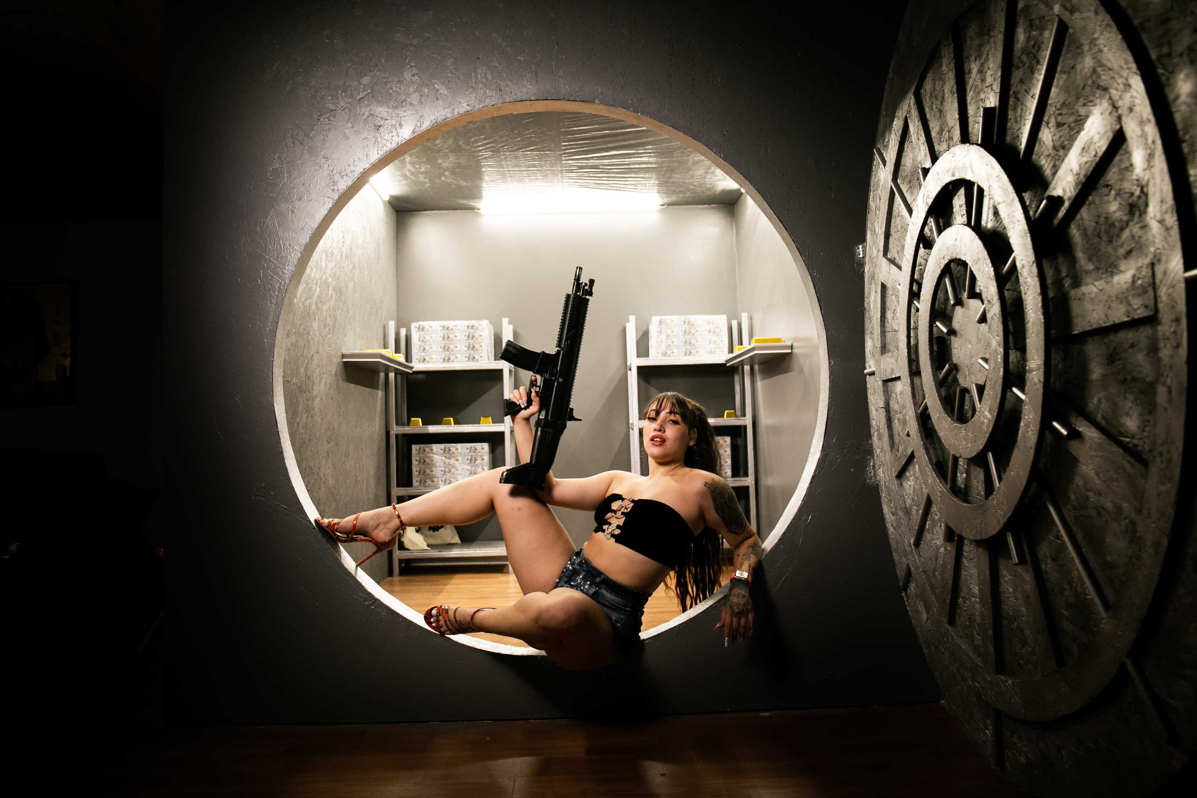 A woman in a black top and denim shorts poses while holding a rifle inside a circular cutout in a wall.