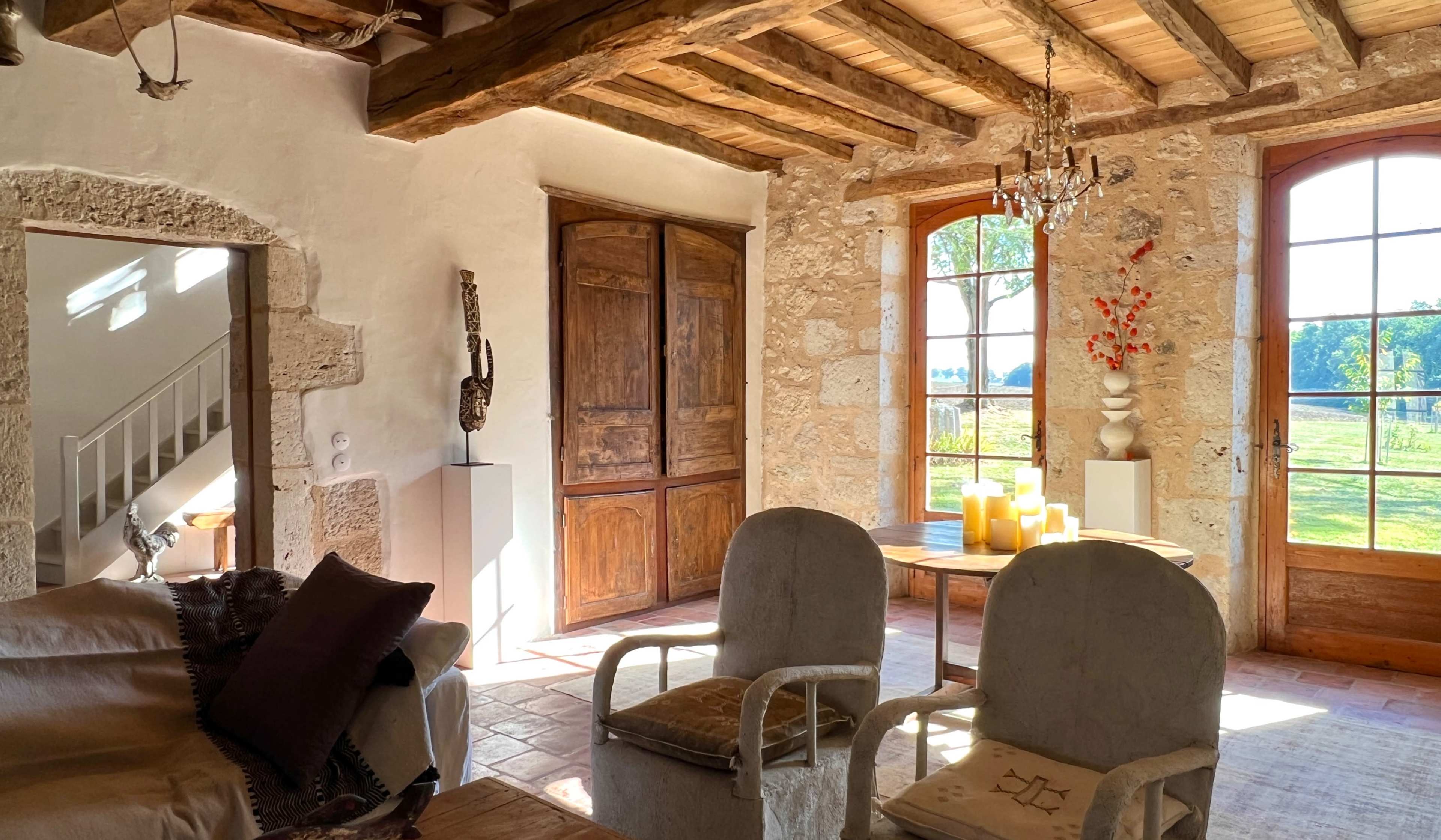 A sunlit living room features rustic wooden beams, stone walls, and a wooden cabinet, with two armchairs facing a table adorned with candles and a view of the outdoors through large windows.