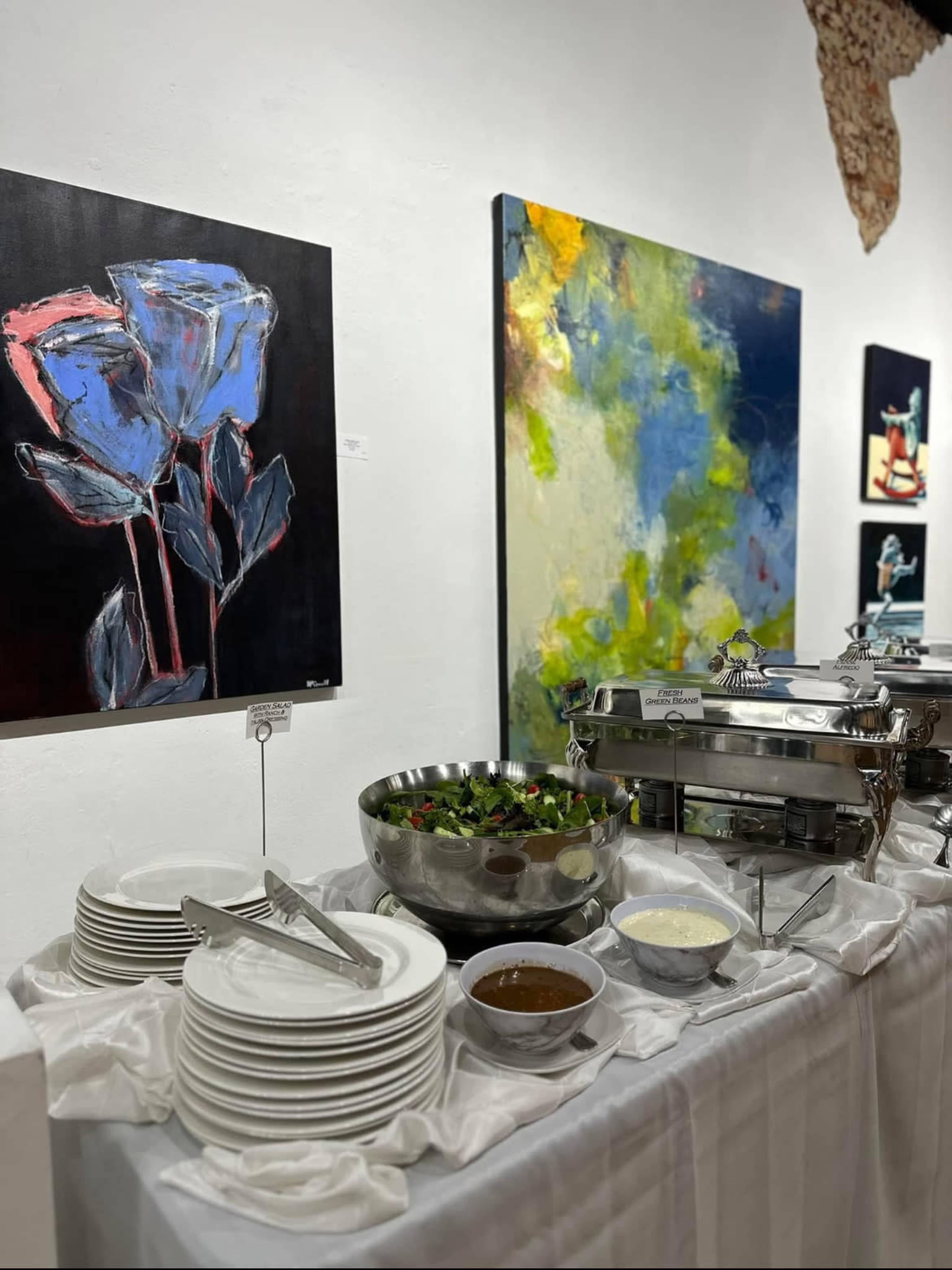The image shows a buffet table adorned with plates, bowls of salad and sauce, and colorful abstract paintings on the walls.