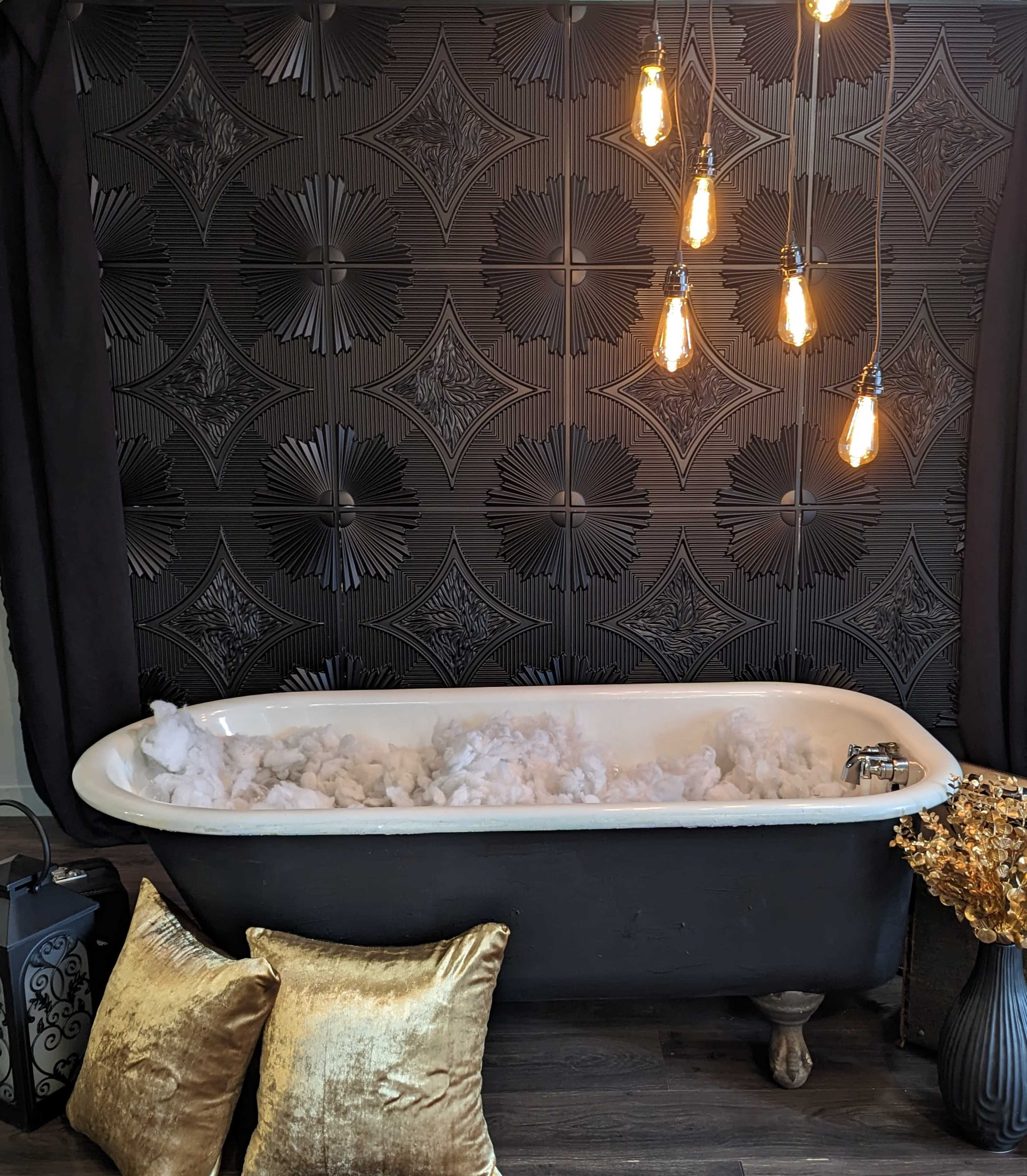A vintage bathtub filled with white fluff sits against a patterned black wall, illuminated by hanging light bulbs, surrounded by decorative pillows and a plant.