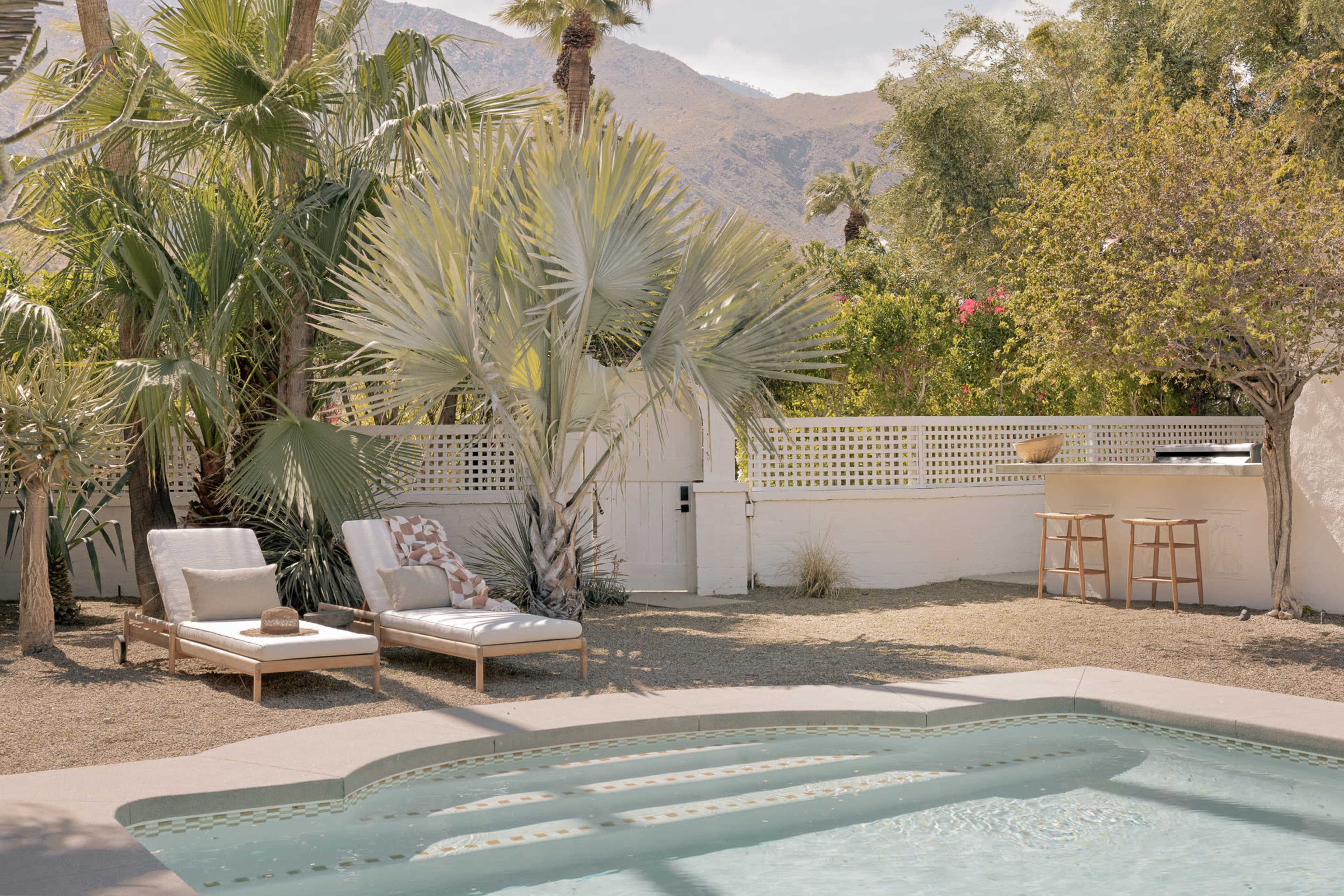 A swimming pool is surrounded by palm trees and lounge chairs in a backyard with a mountain backdrop.