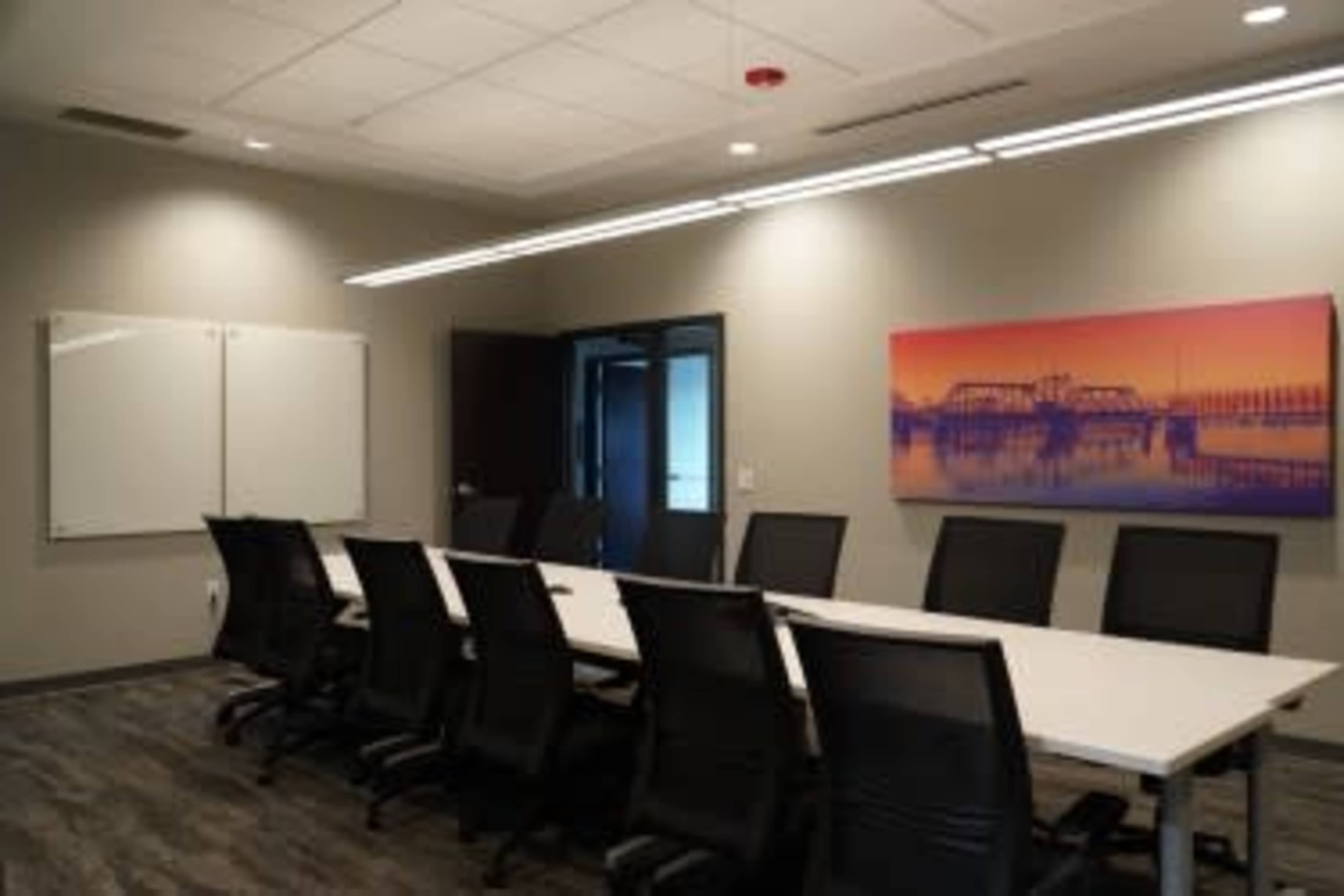 The image shows a modern conference room featuring a long table surrounded by black chairs, with a large wall-mounted artwork and two whiteboards.