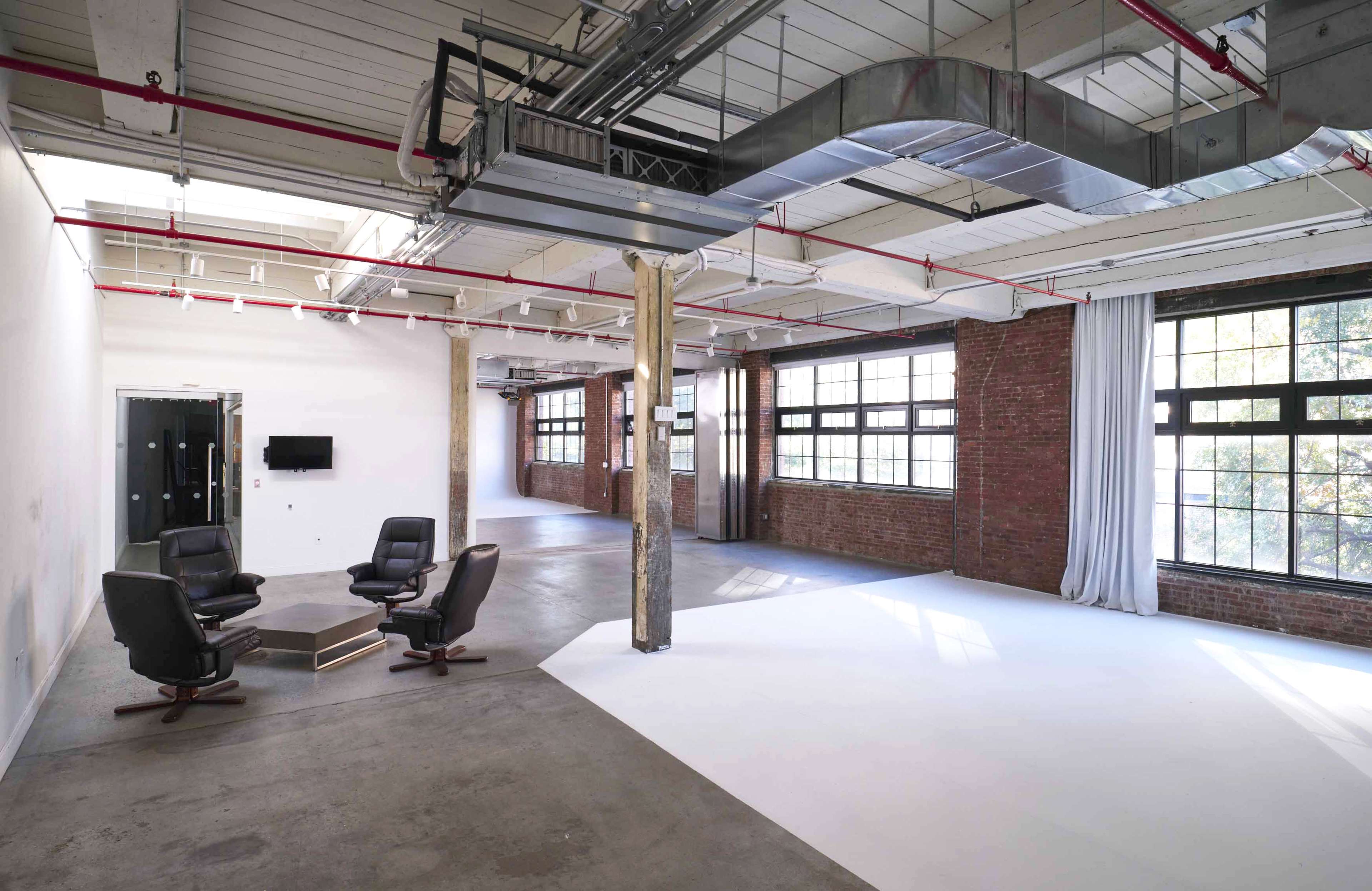 An industrial-style interior space with concrete floors, exposed beams, and large windows, featuring a small seating area with two black armchairs and a wooden coffee table.
