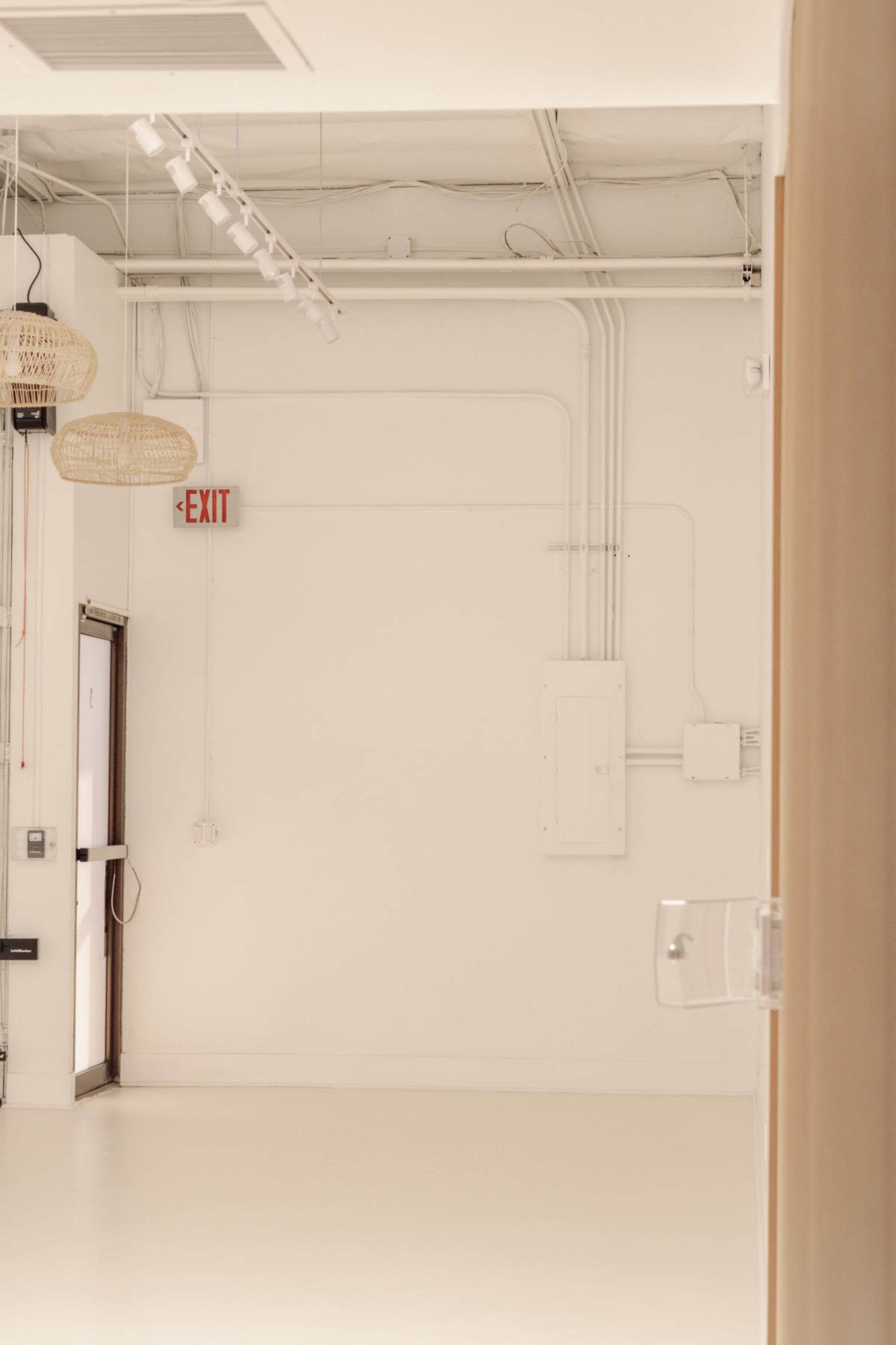 The image shows a minimalist interior space with white walls, overhead lighting, and an exit sign near a door.