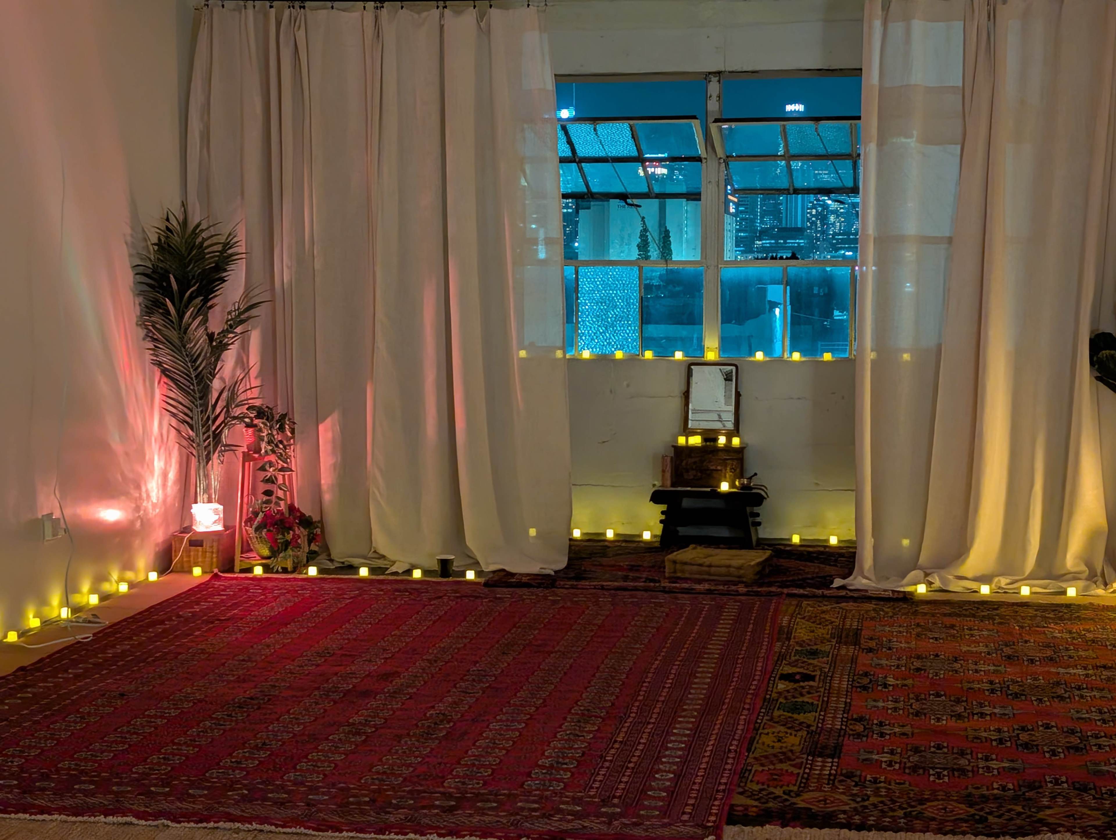 The image shows a dimly lit room with a patterned rug, decorated with candles arranged along the walls and a window revealing a city skyline.