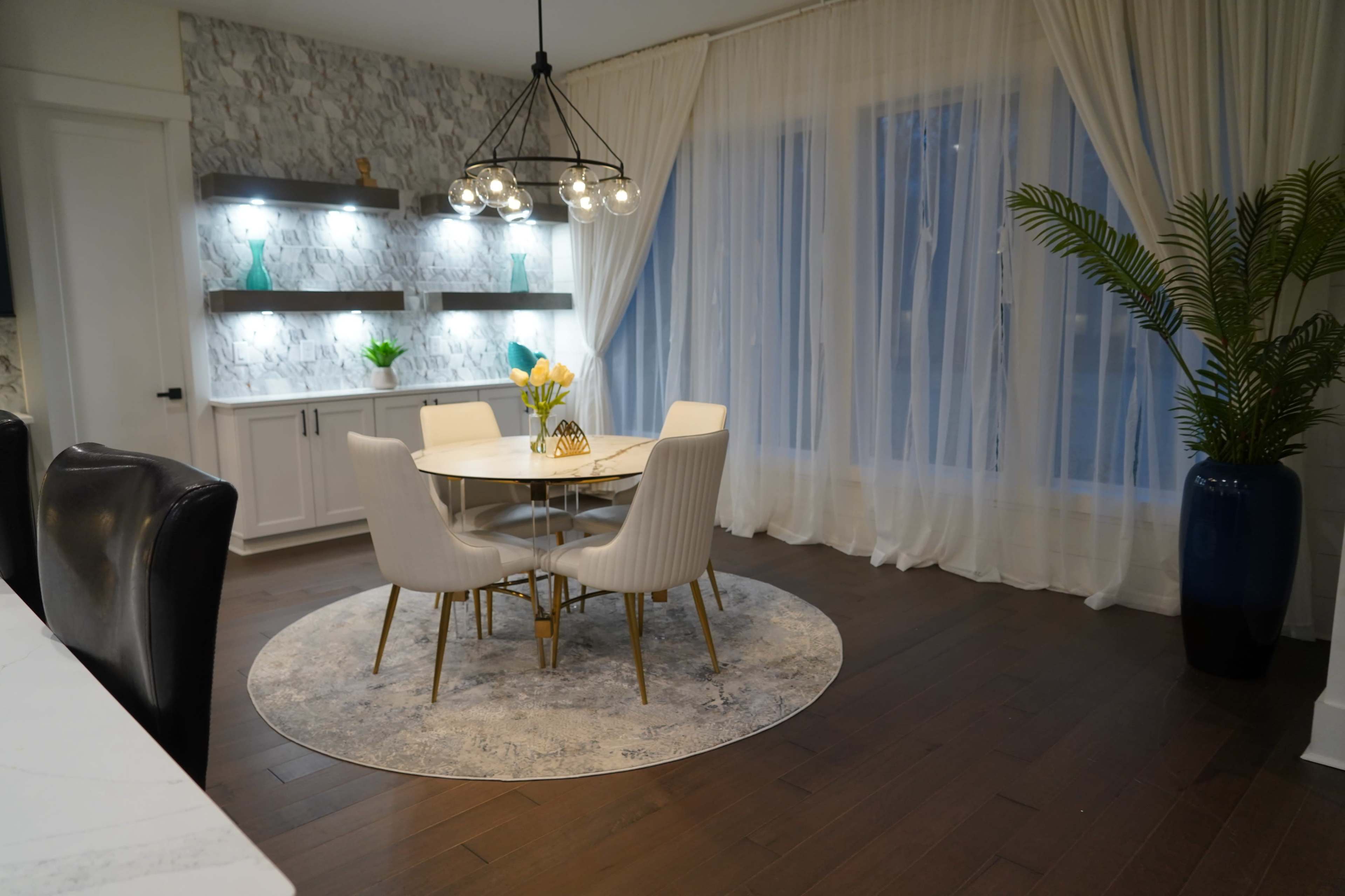 The image shows a modern dining area with a round table surrounded by four upholstered chairs, illuminated by a chandelier, and accented by a large window draped with sheer curtains.