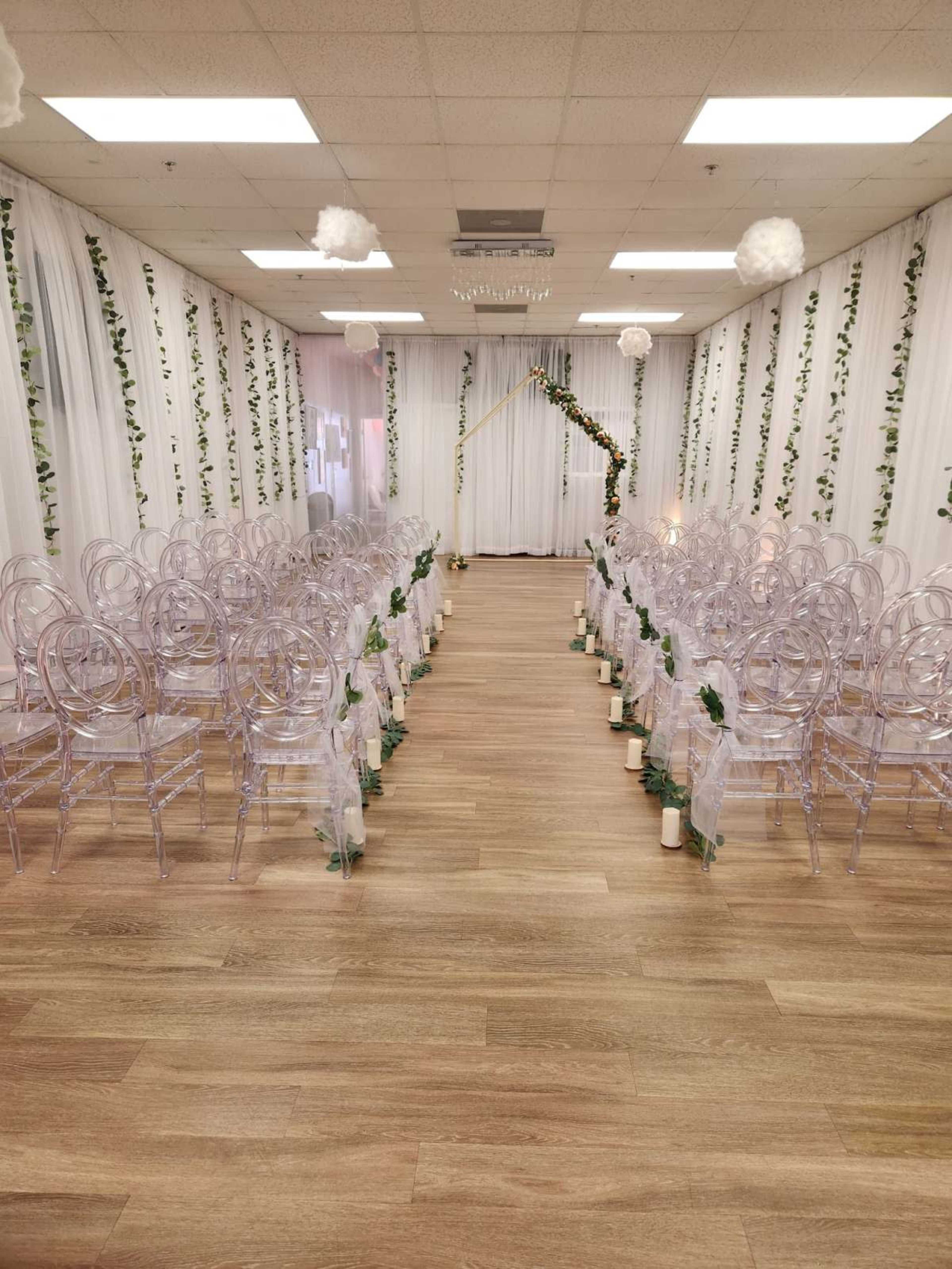 A wedding ceremony setup with clear plastic chairs arranged in two rows facing an altar adorned with greenery and draped fabric.