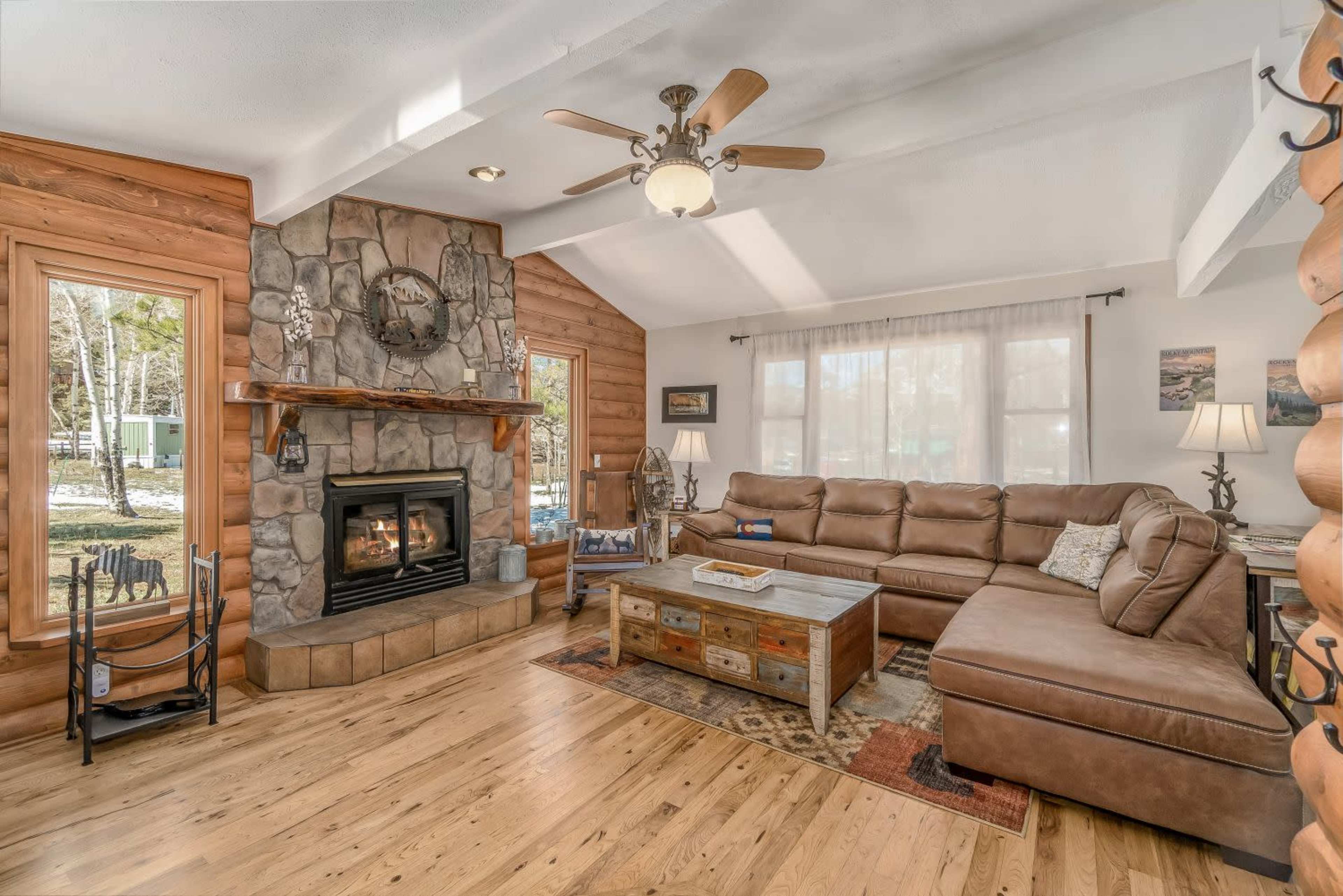 A cozy living room with a stone fireplace, large windows, a sectional sofa, and wooden flooring.