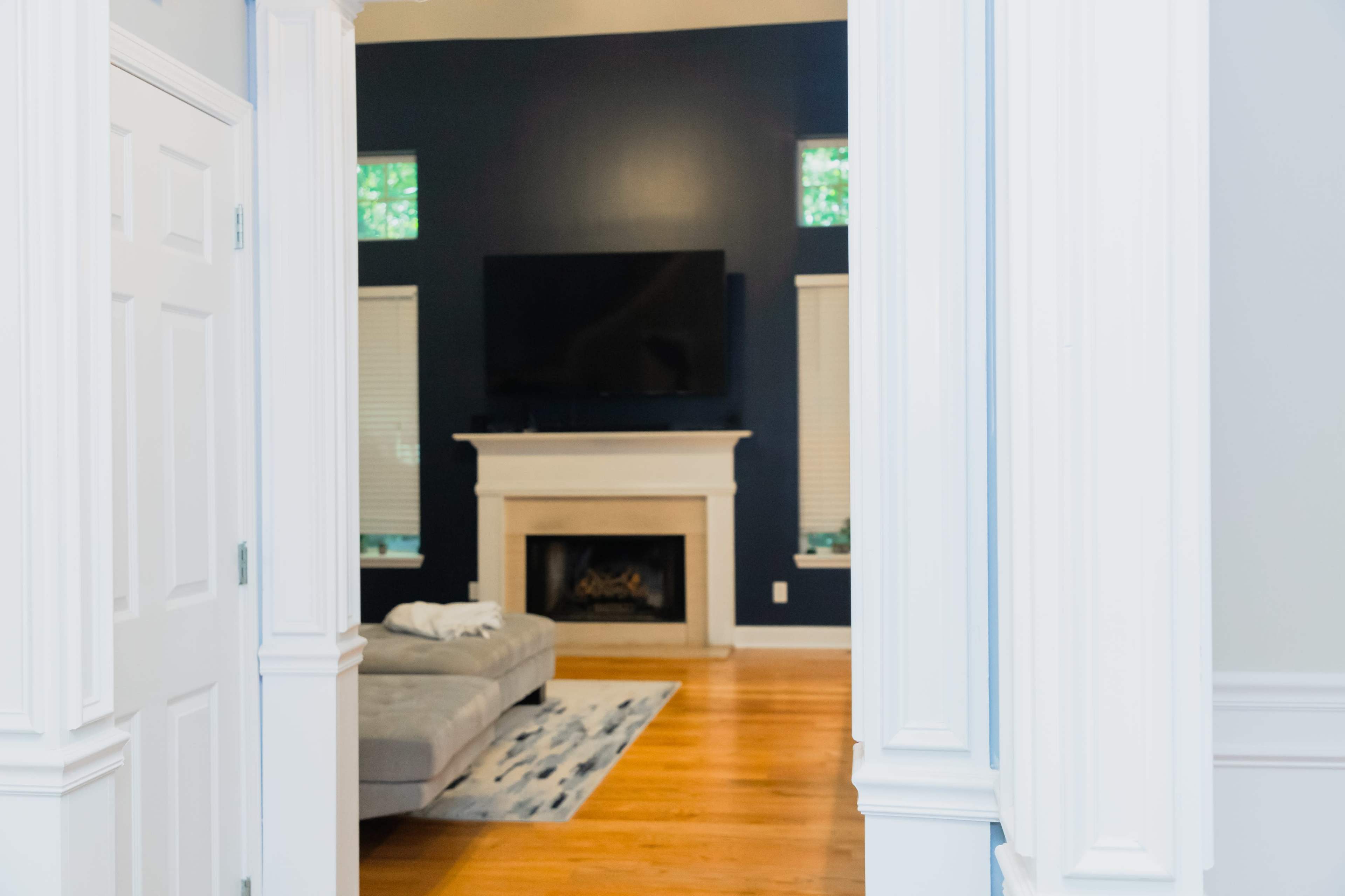 A modern living room features a grey sofa, a fireplace, and a wall-mounted television, framed by white columns in the foreground.