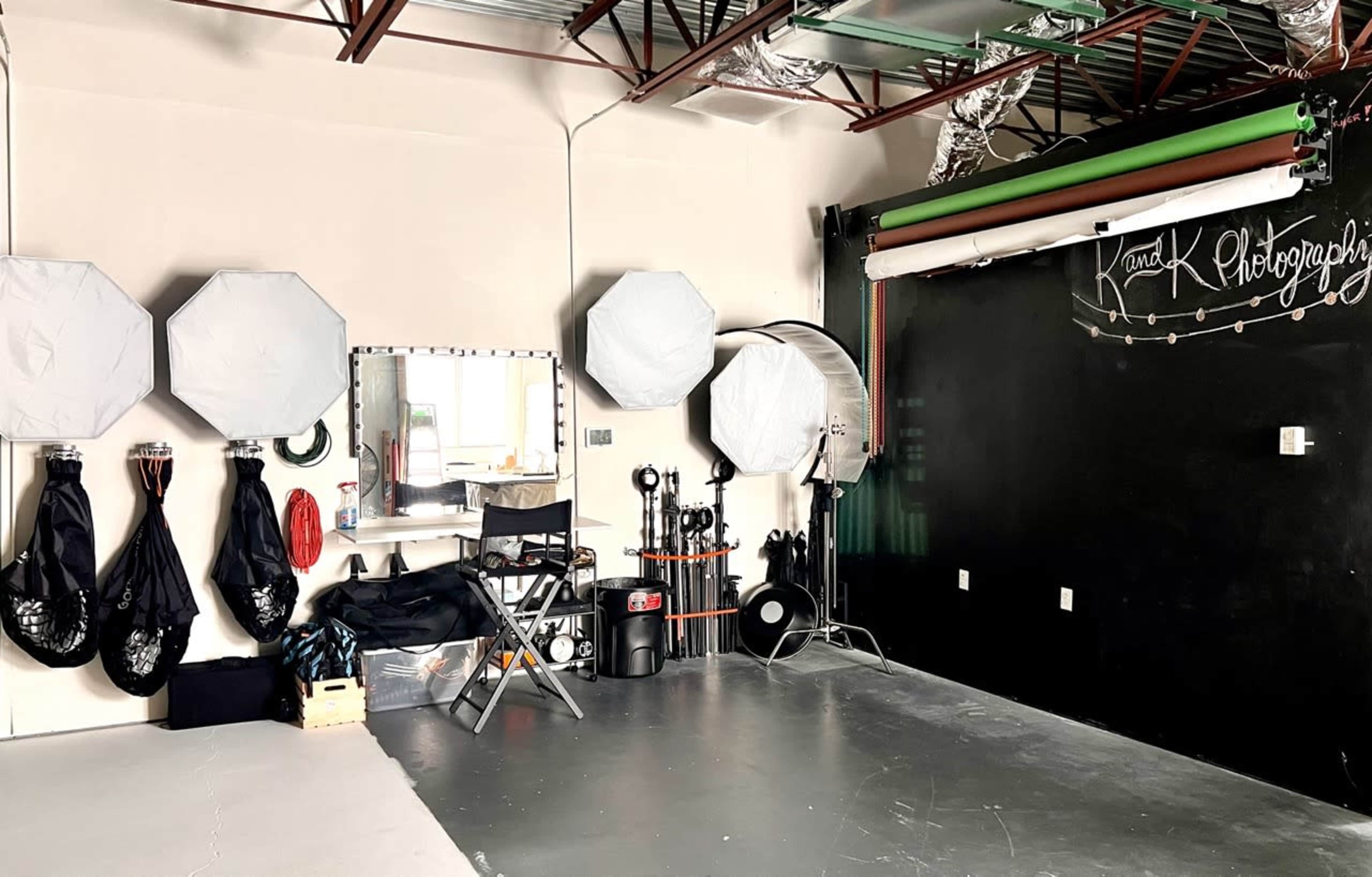 A photography studio featuring a mirror with lights, multiple softbox lights, a director's chair, and a black wall with the text "K and K Photography."