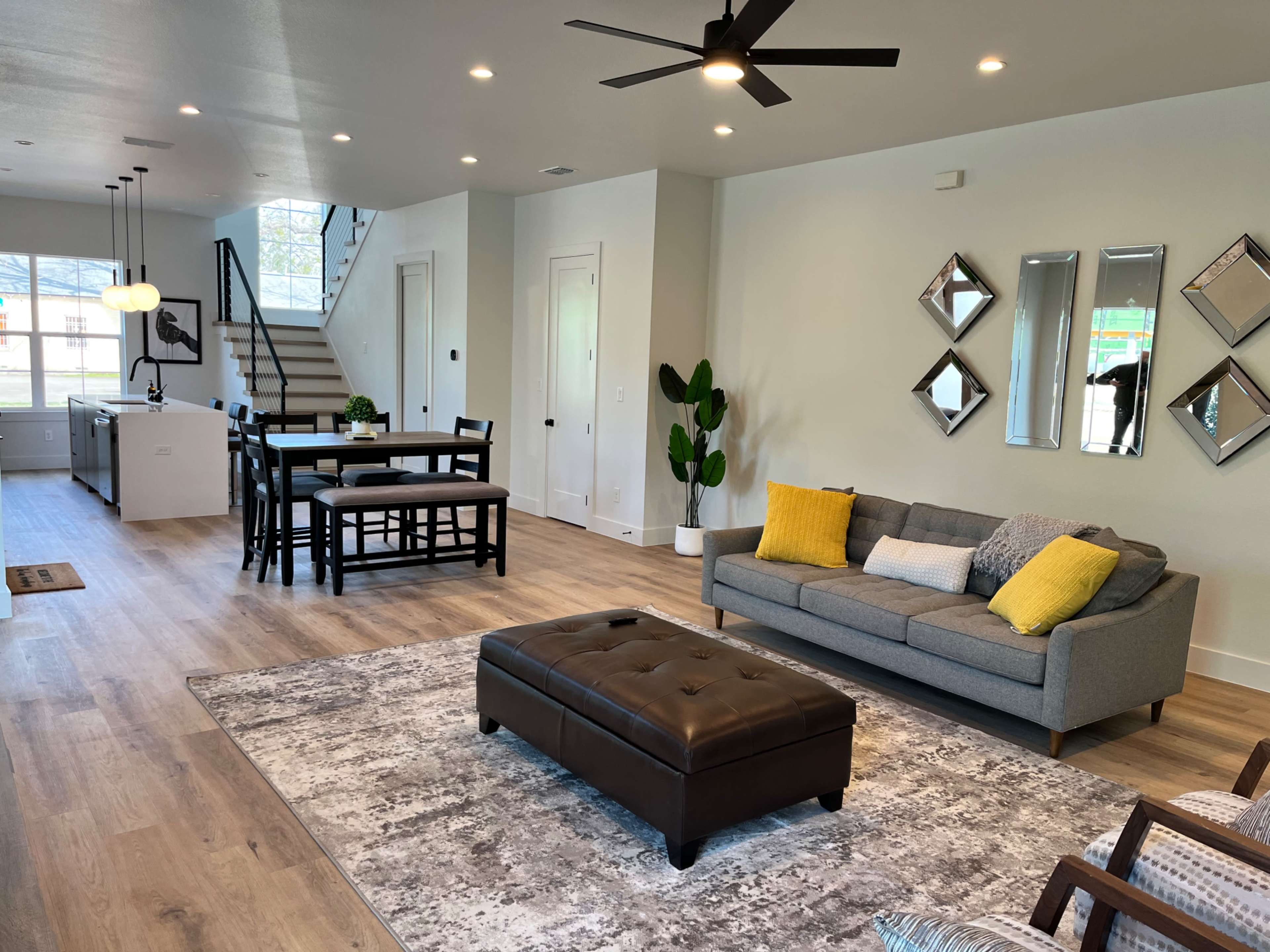 A modern living space with a gray sofa, a black ottoman, a dining table, and a staircase, all illuminated by natural light.