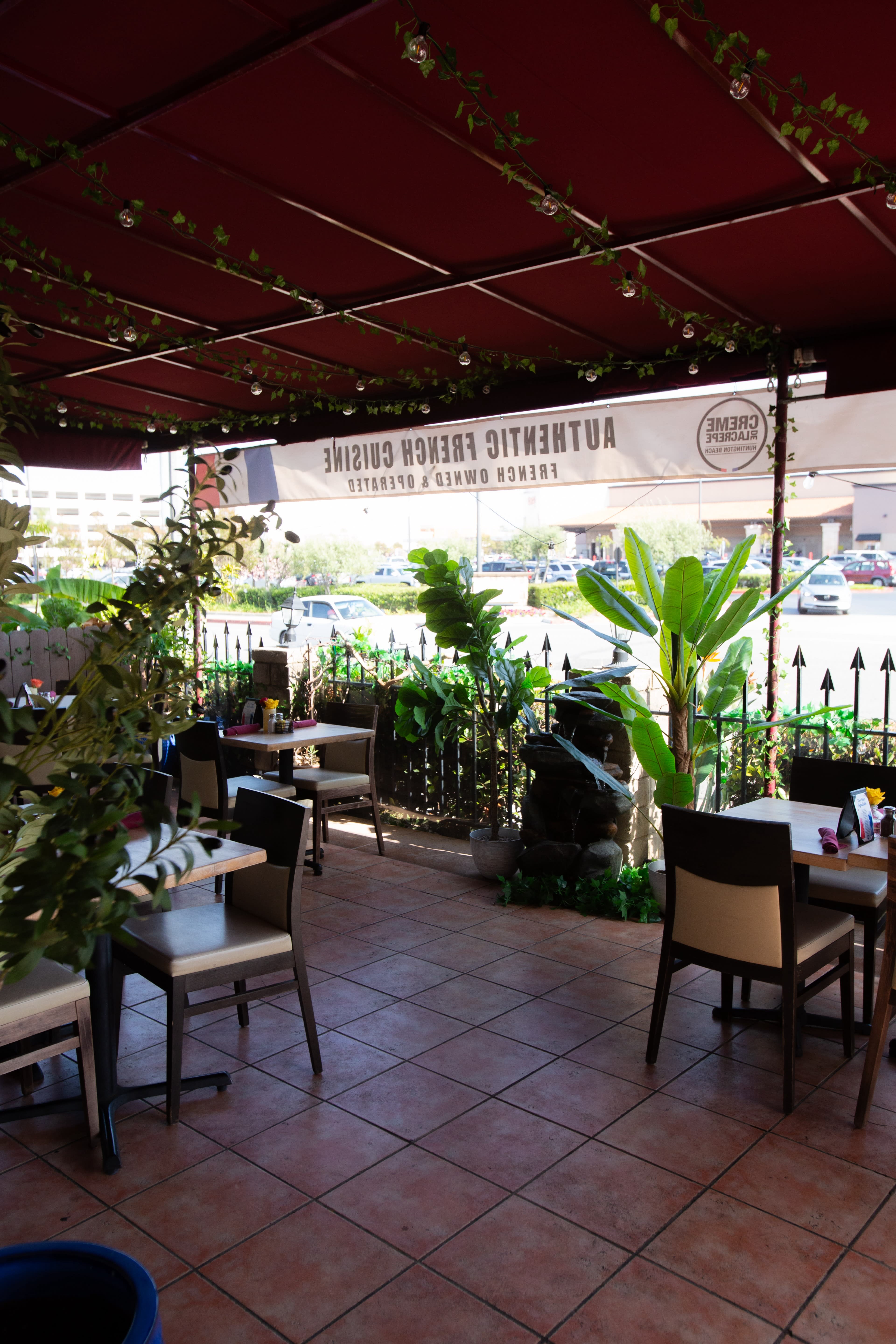 The image shows a covered outdoor seating area of a restaurant with tables and chairs arranged around potted plants and a decorative water feature.