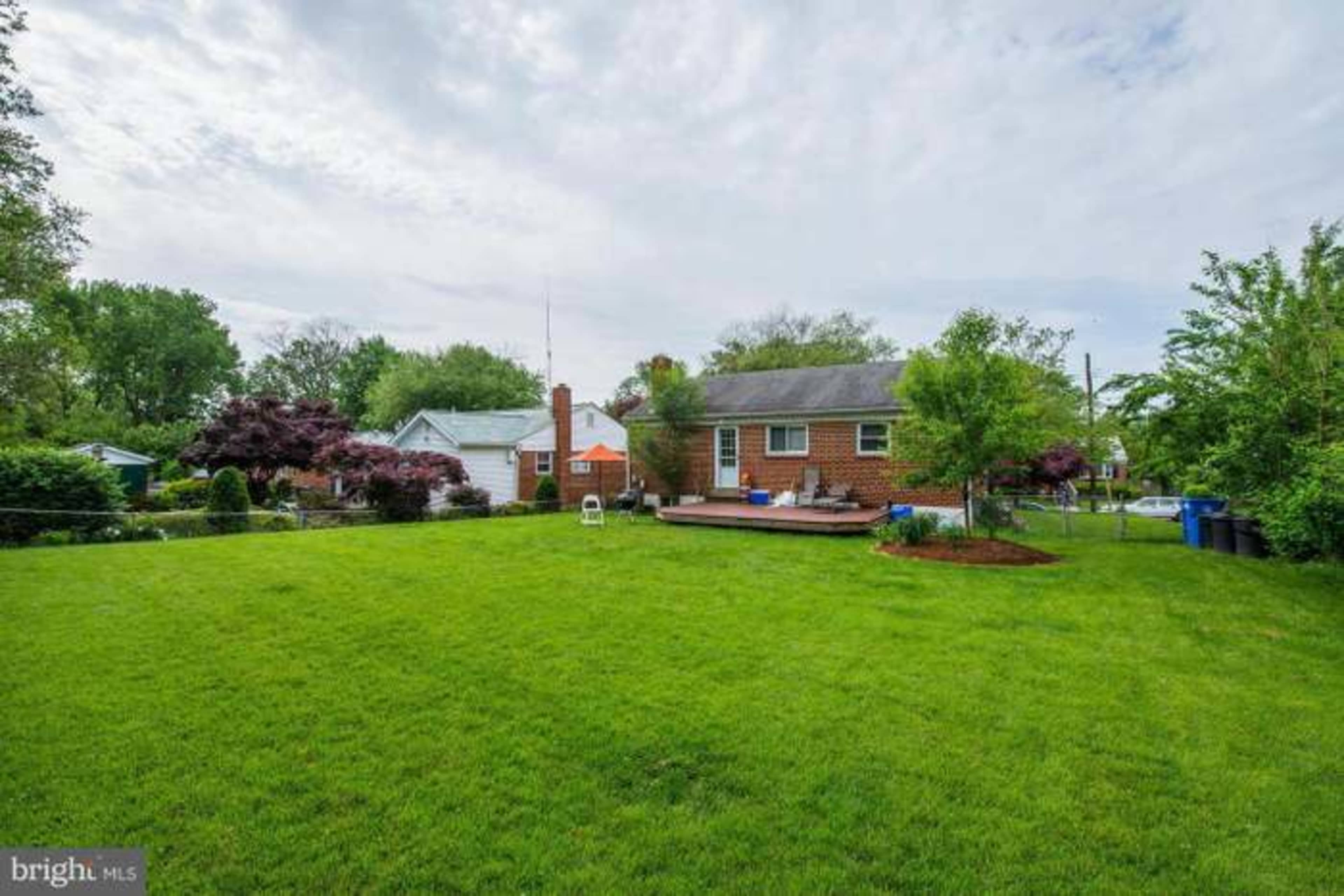A single-story brick house with a deck is surrounded by a spacious lawn and several trees in a residential neighborhood.