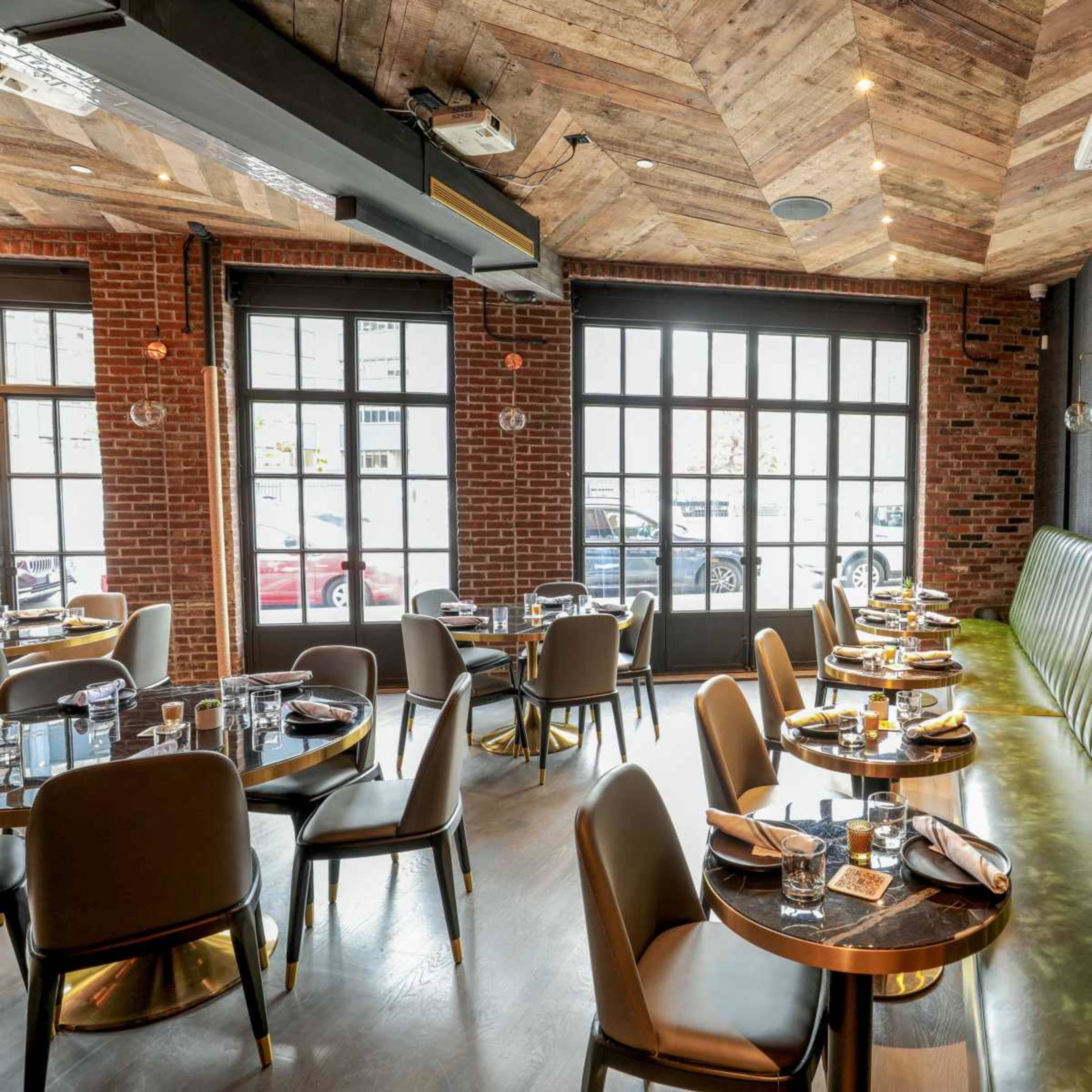 The interior of a modern restaurant features wooden ceilings, brick walls, and round tables set with dinnerware.
