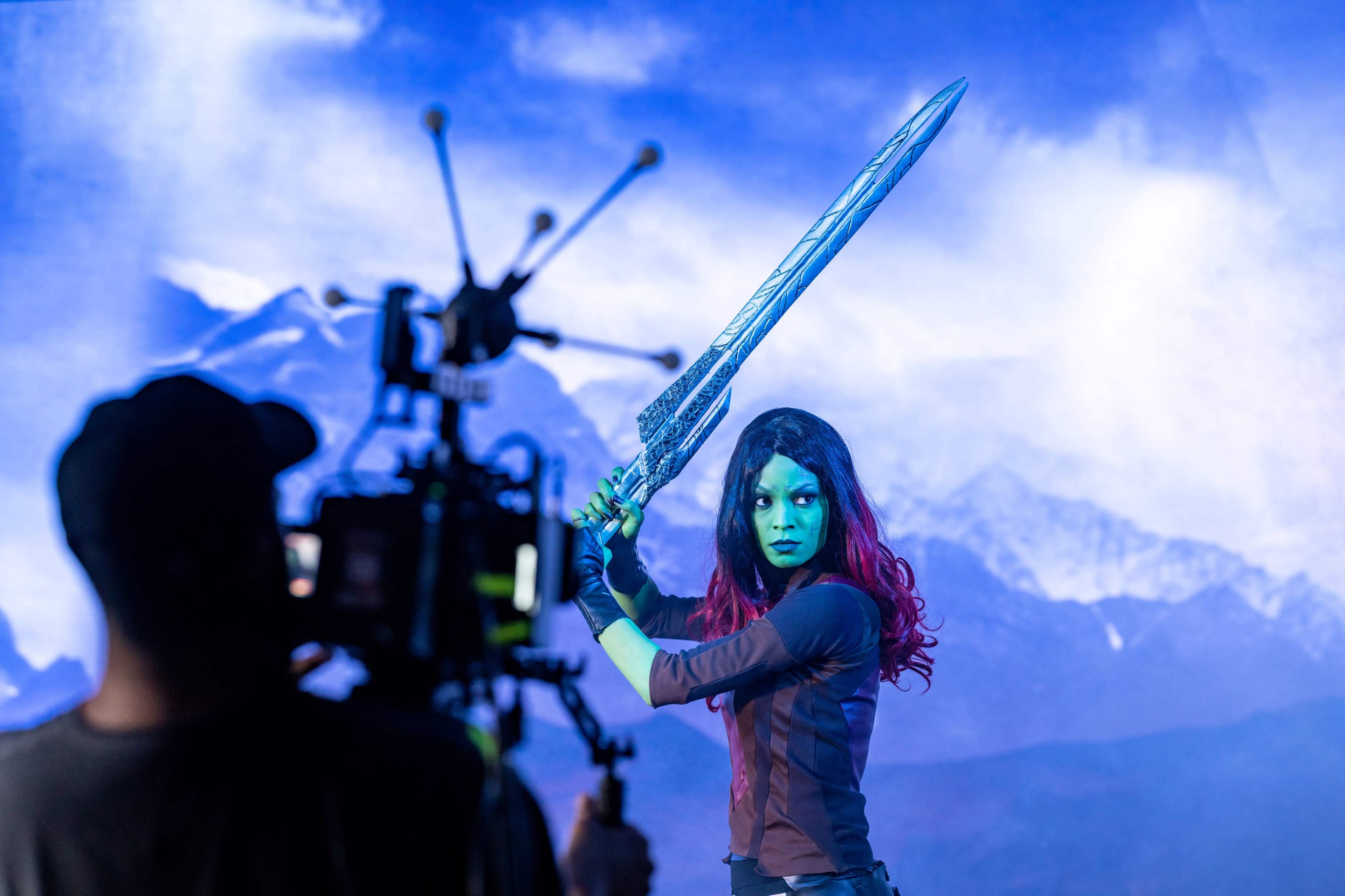 A person with green skin holds a large sword while posing in front of a mountainous backdrop and a film crew.