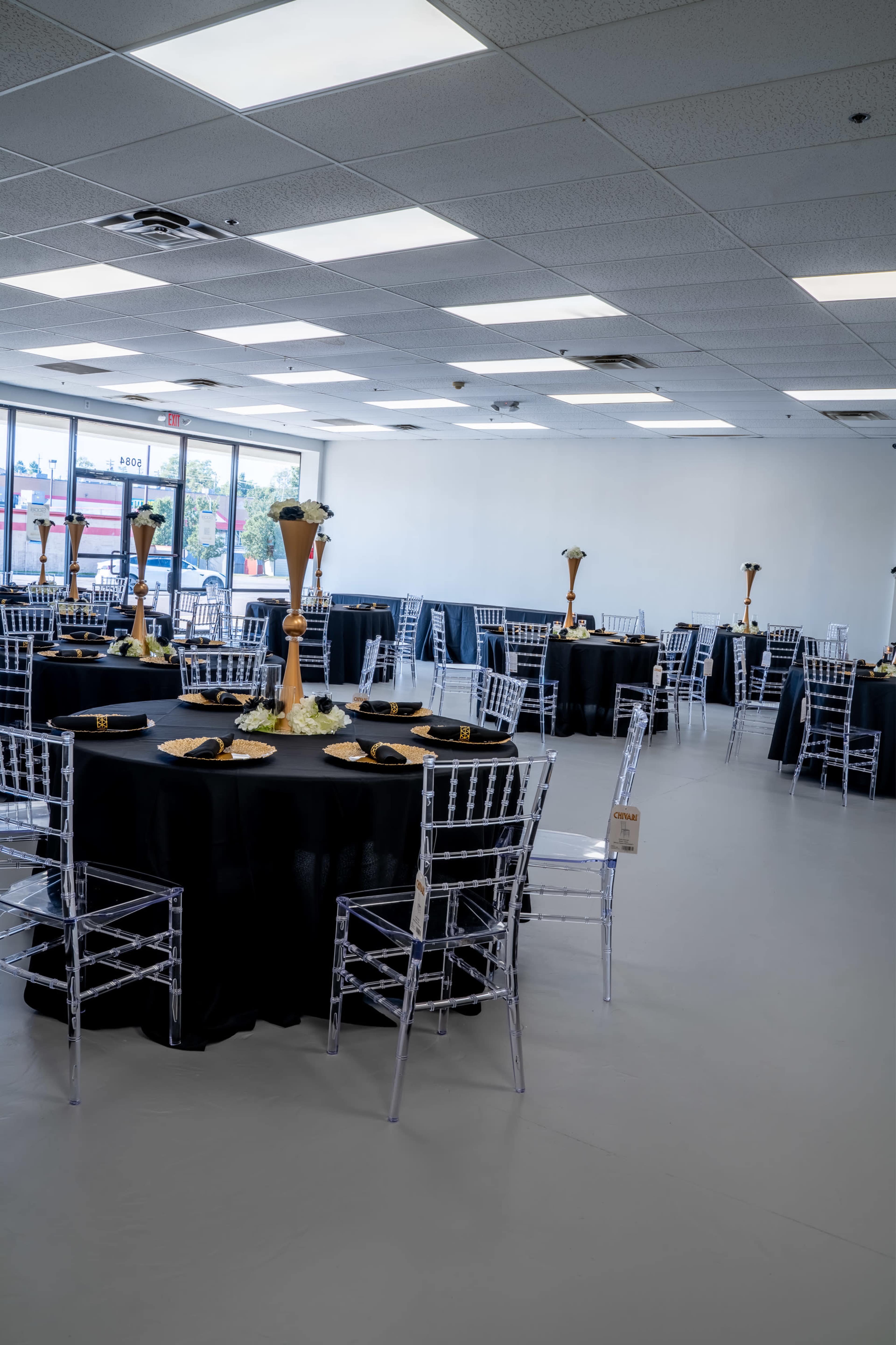 The image shows a spacious event hall with several round tables covered in black tablecloths, each adorned with tall centerpieces and white floral arrangements.
