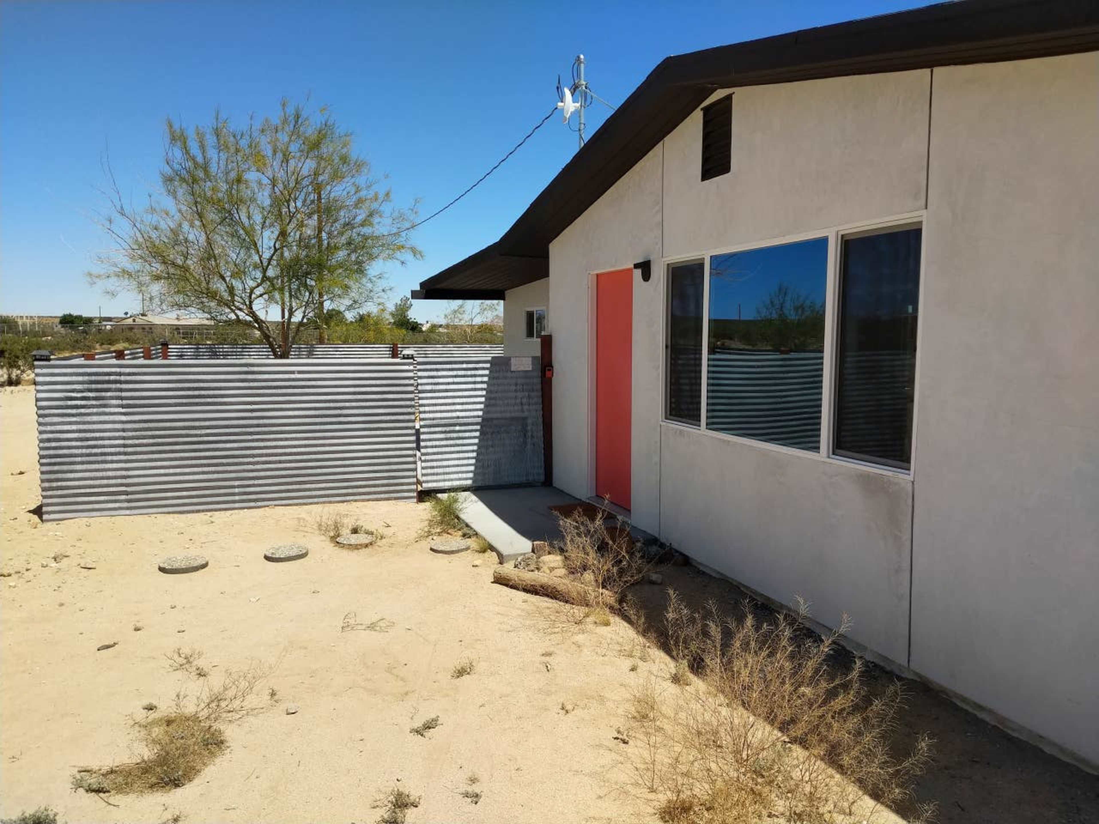 A modern house with a bright orange door is situated in a sandy landscape, surrounded by sparse vegetation and a corrugated metal fence.