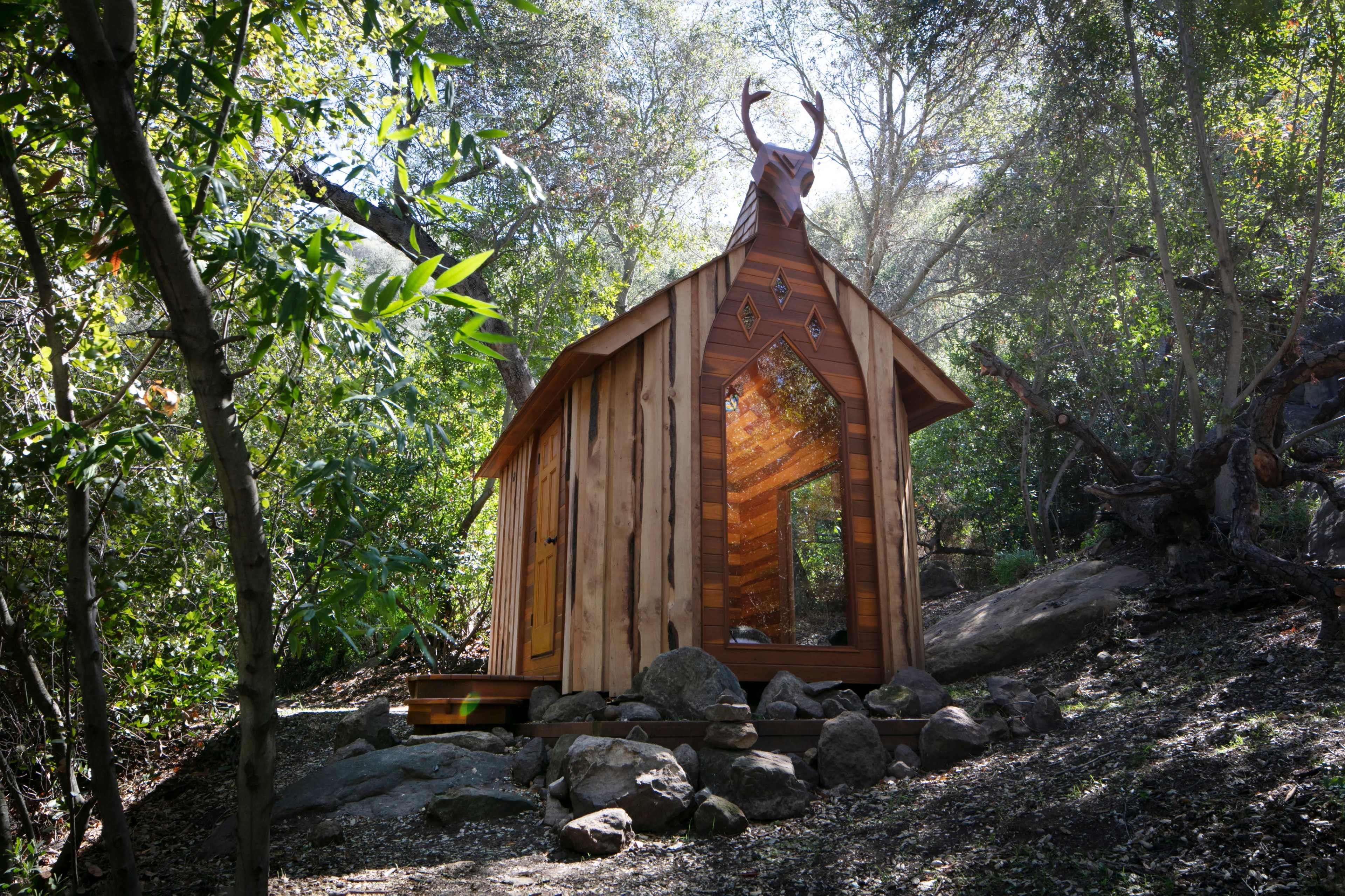 A small wooden cabin with a peaked roof and deer antlers on top is nestled among trees and rocks in a forested area.