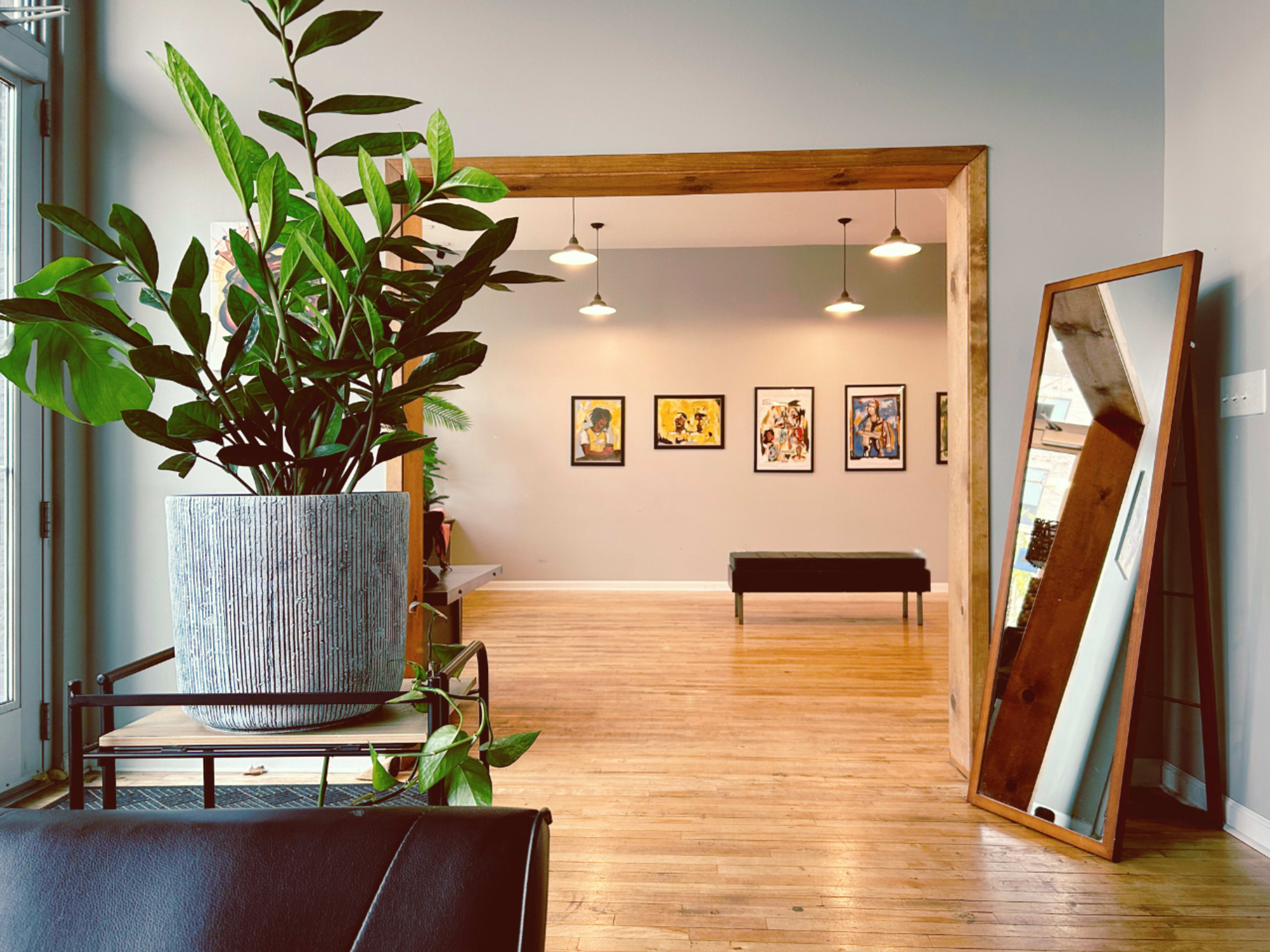 An indoor space features a potted plant, a large mirror, and framed artwork on the wall opposite a bench.
