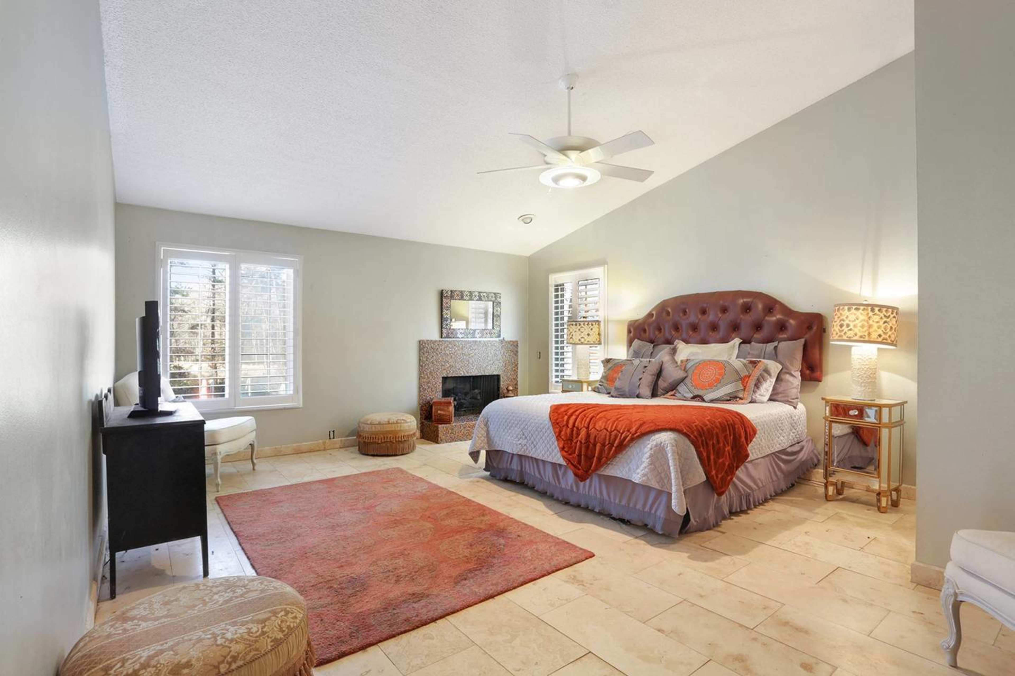 The spacious bedroom features a large bed with a tufted headboard, two accent lamps, a fireplace, and a rug on tiled flooring.