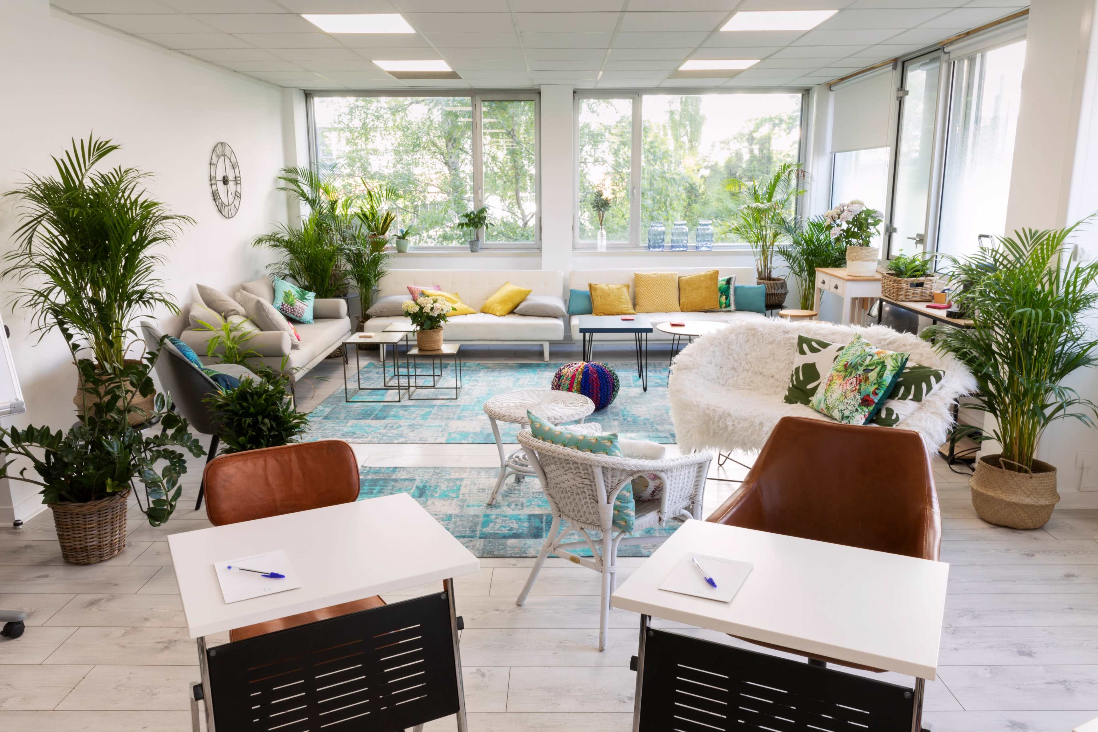 A bright, modern workspace featuring comfortable seating areas, green plants, and two desks with pens on them.