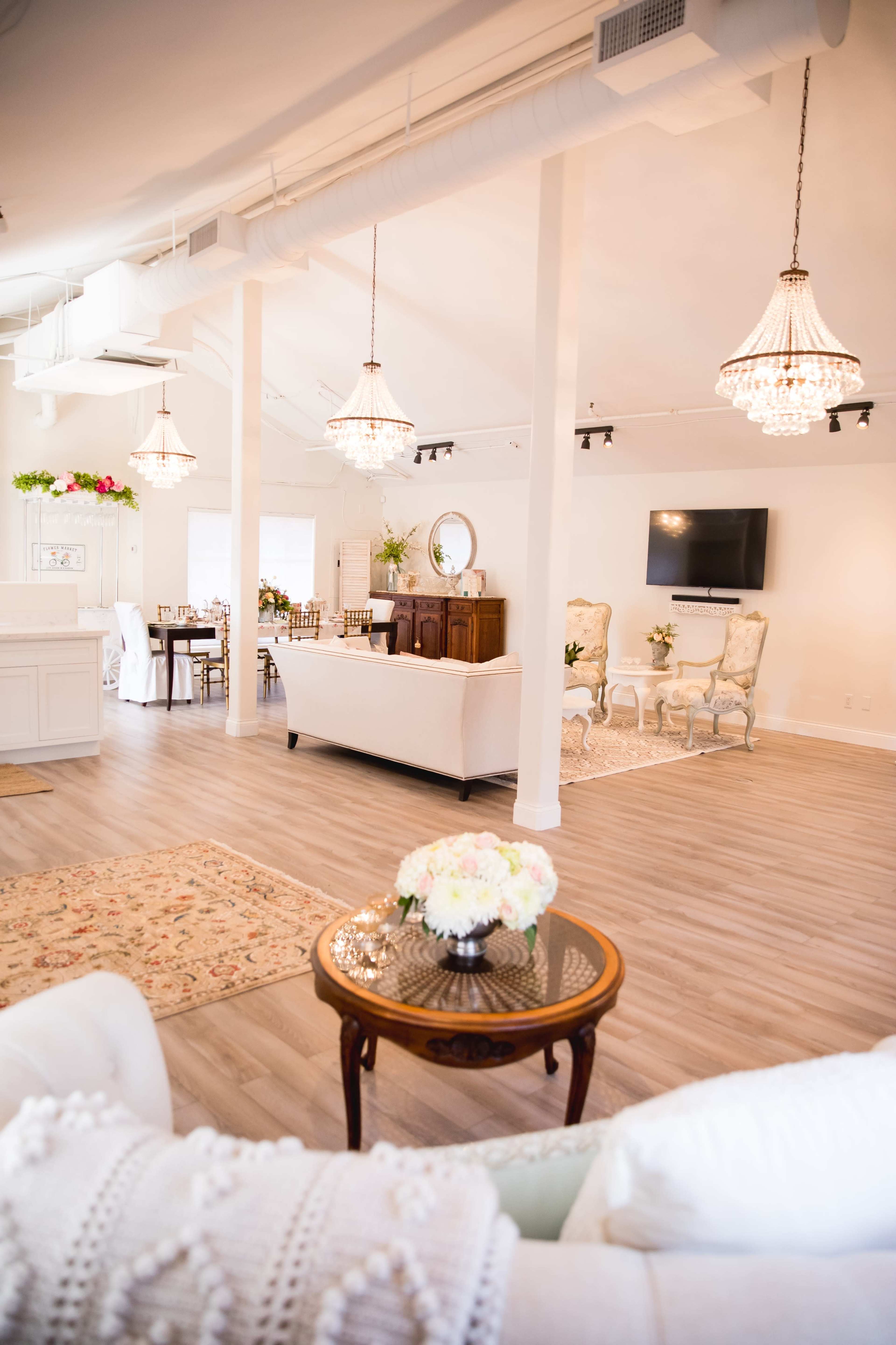 An elegantly decorated event space with a light-colored sofa, crystal chandeliers, dining tables, and a large television mounted on the wall.