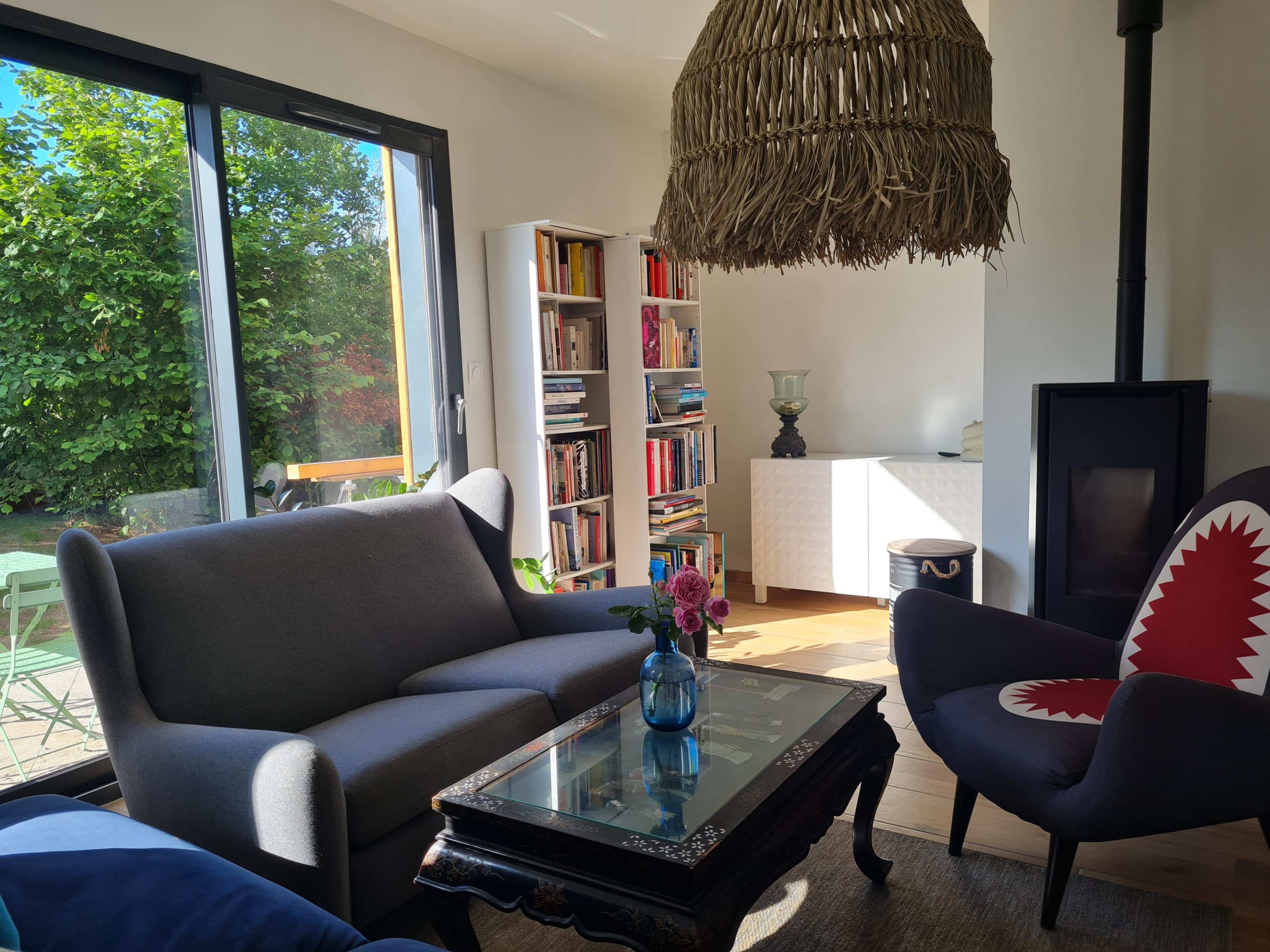 A cozy living room with a large window, showcasing a bright outdoor space, a bookshelf filled with books, and two distinct chairs arranged around a coffee table.