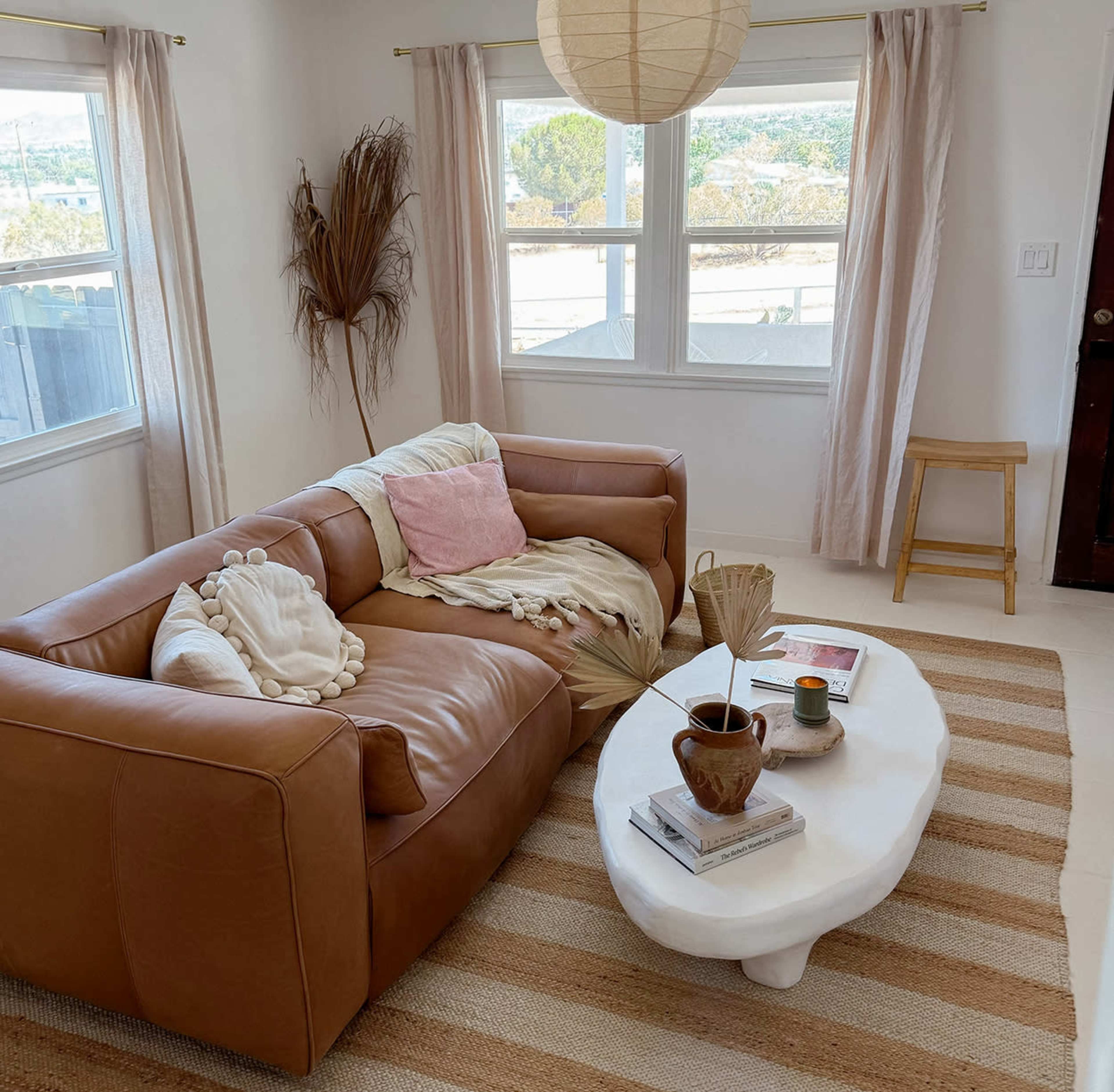 A cozy living room featuring a brown leather sofa, a white coffee table, and natural light streaming through sheer curtains.