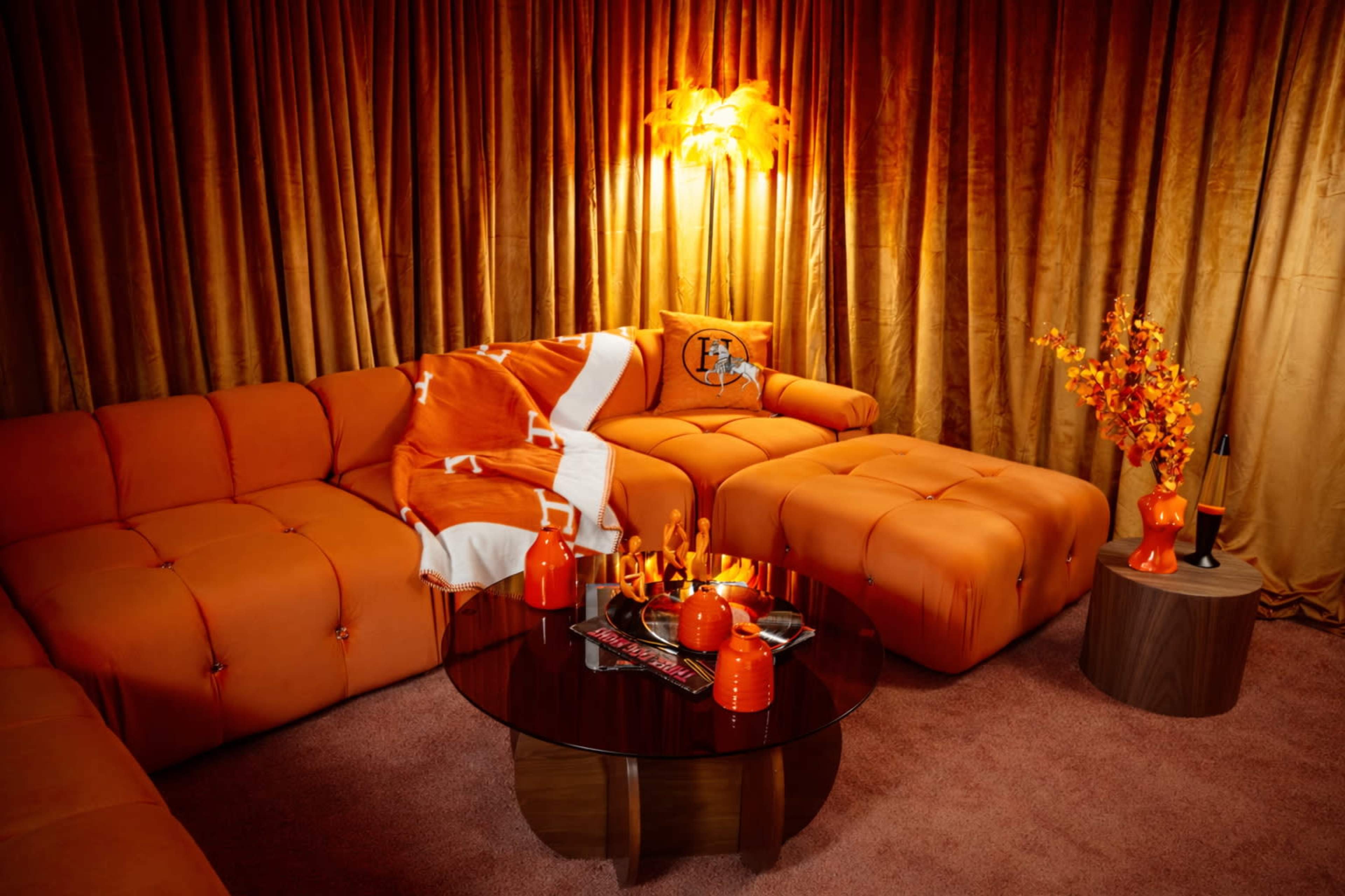 The image depicts a warmly colored living room featuring an orange sectional sofa, a round coffee table with decorative items, and an accent chair, all surrounded by matching drapes.