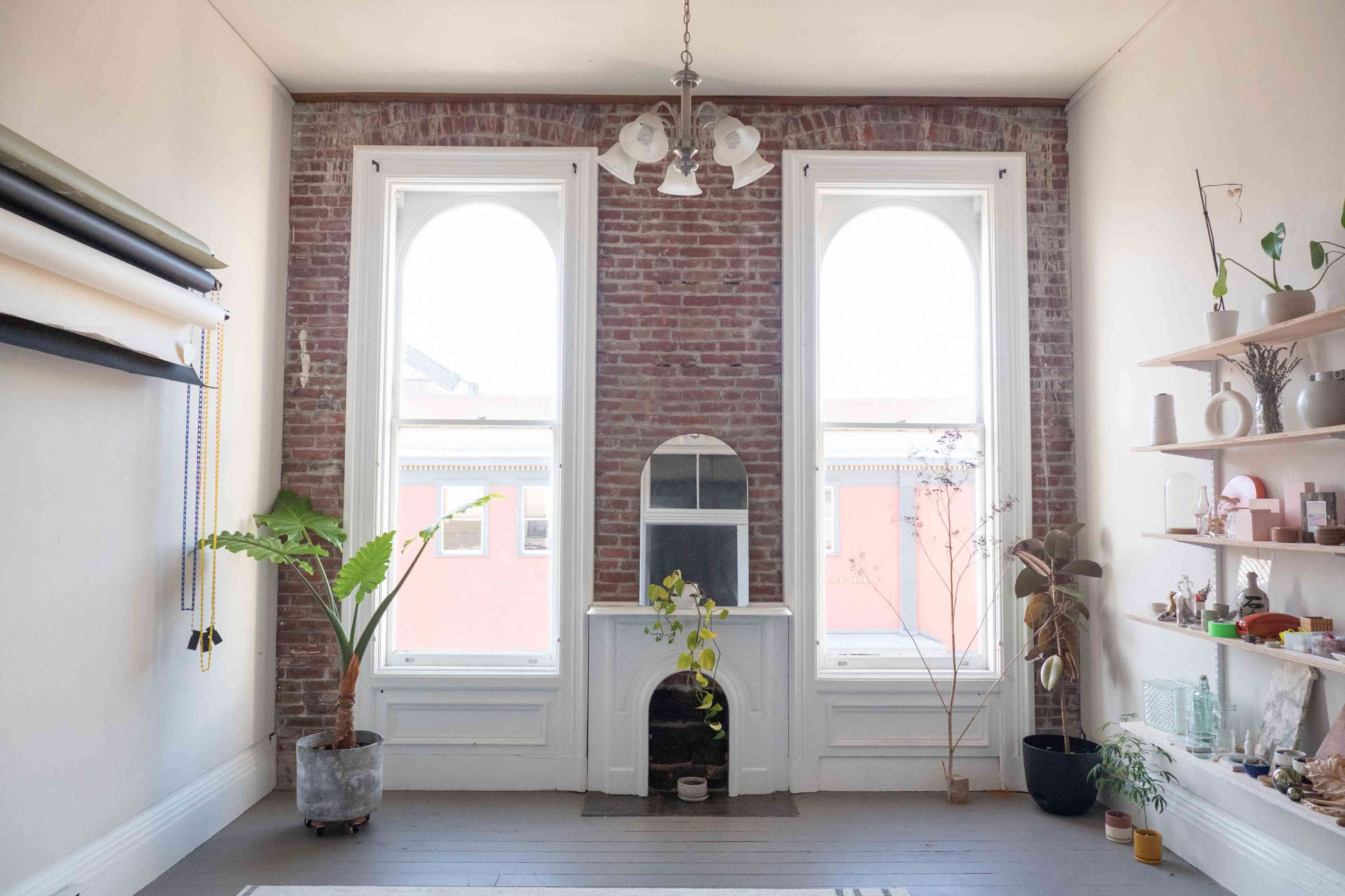 The image shows a minimalistic room with exposed brick walls, large arched windows, a mirror above a small fireplace, and various plants and decorative items on shelves.