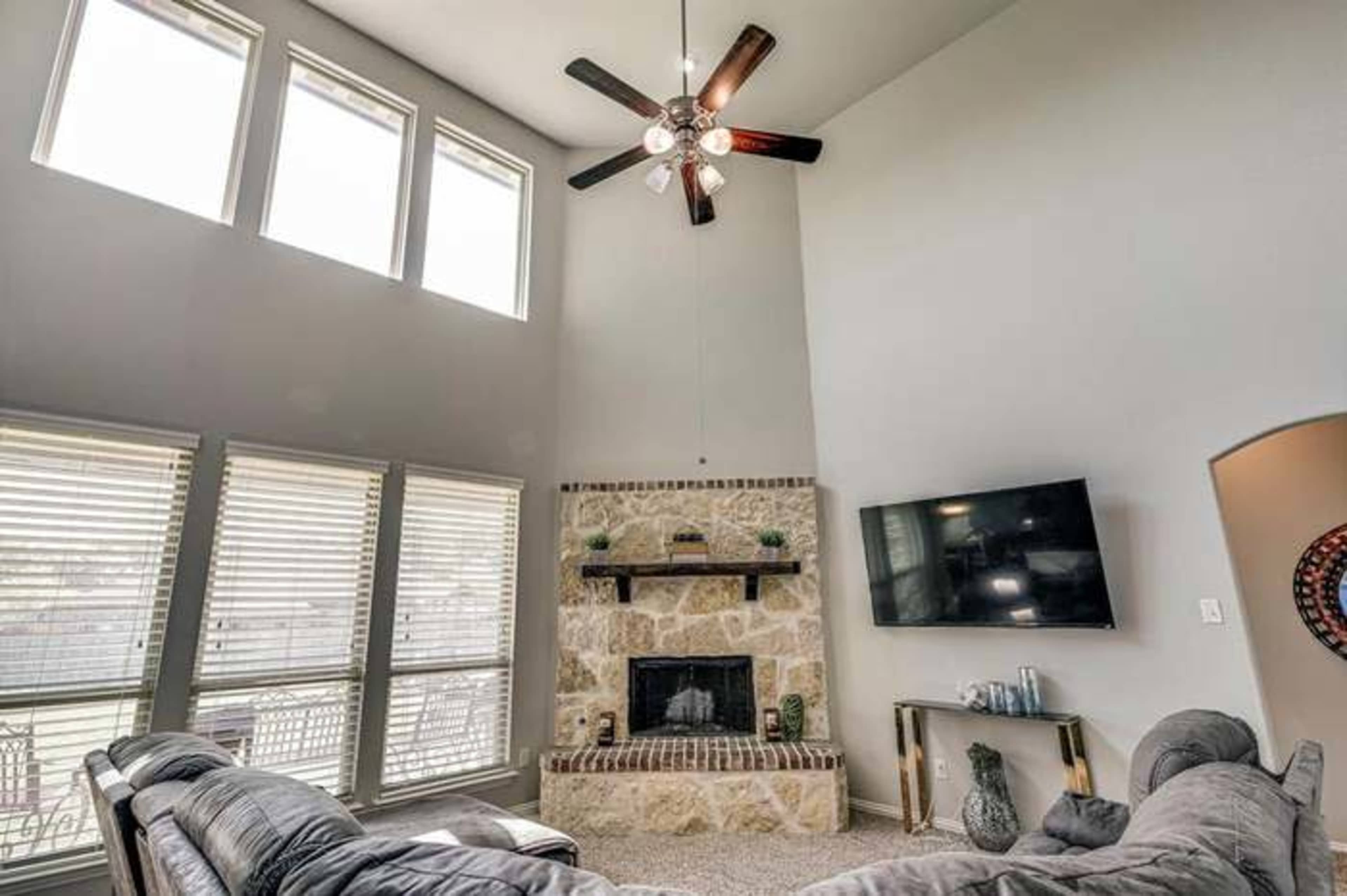 The image shows a spacious living room with a high ceiling, large windows, a stone fireplace, and a wall-mounted television.