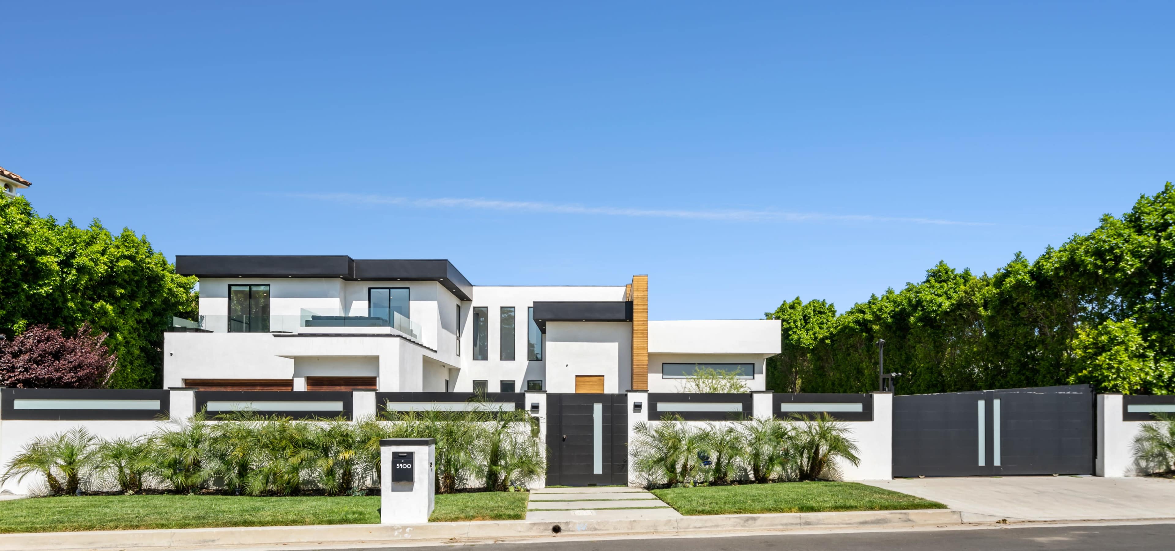 A modern two-story house surrounded by a low wall and landscaped greenery, with a driveway leading to a gated entrance.