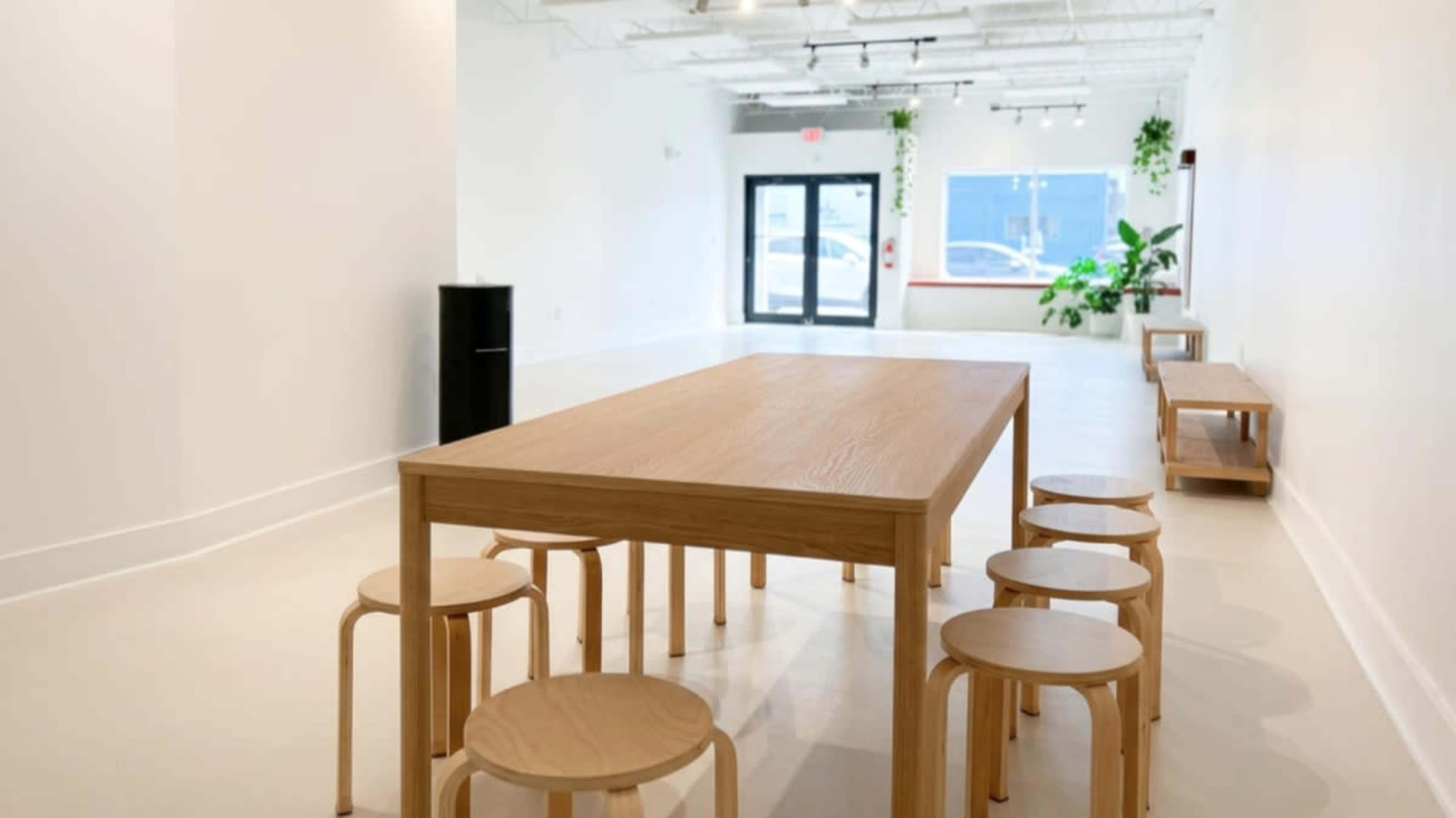 A large wooden table surrounded by several stools is set in a spacious, minimalistic room with white walls and a few indoor plants.