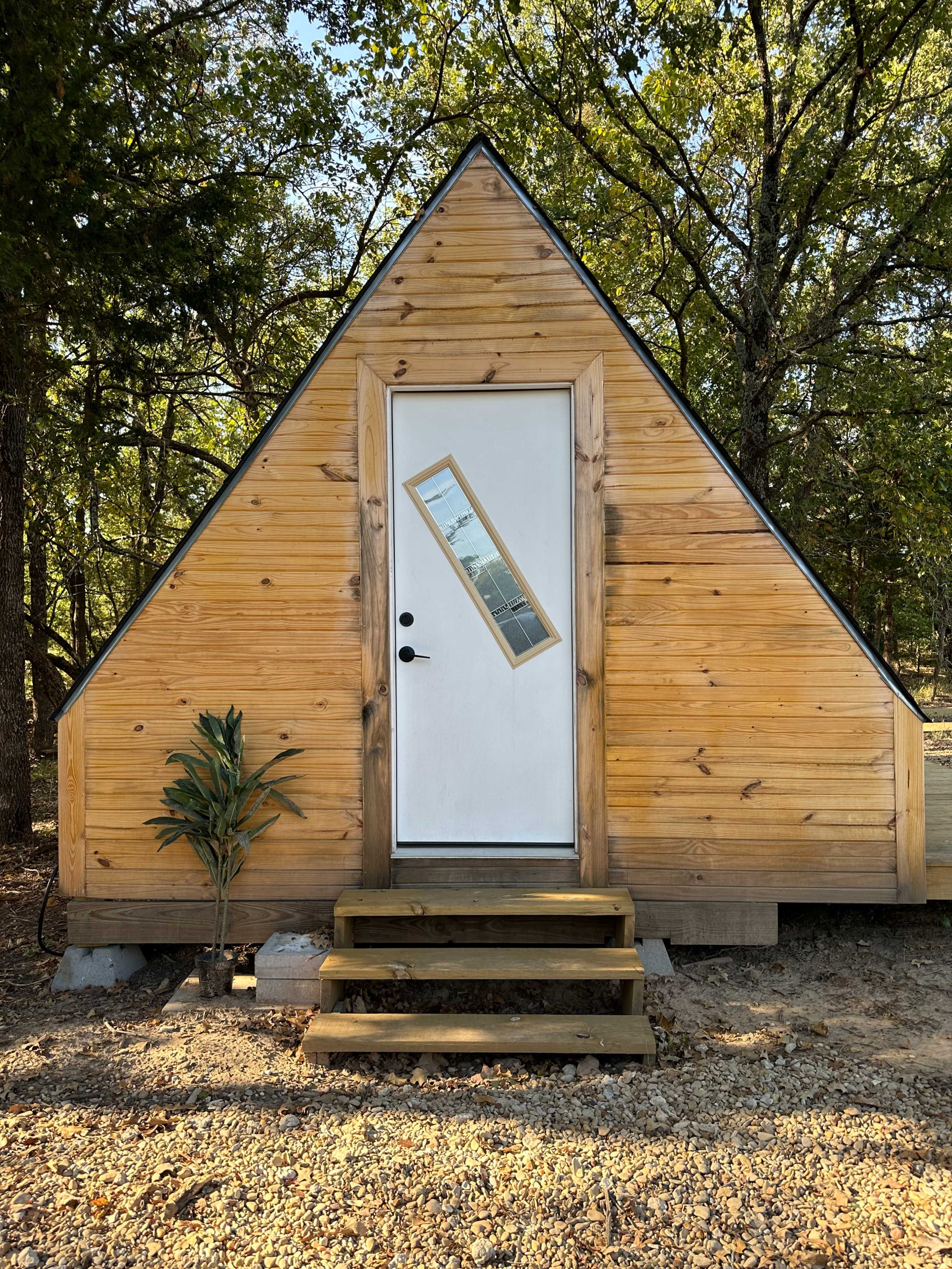 The image shows a small, modern A-frame cabin with a wooden exterior and a distinctive angled roof, featuring a sleek door with a rectangular window.