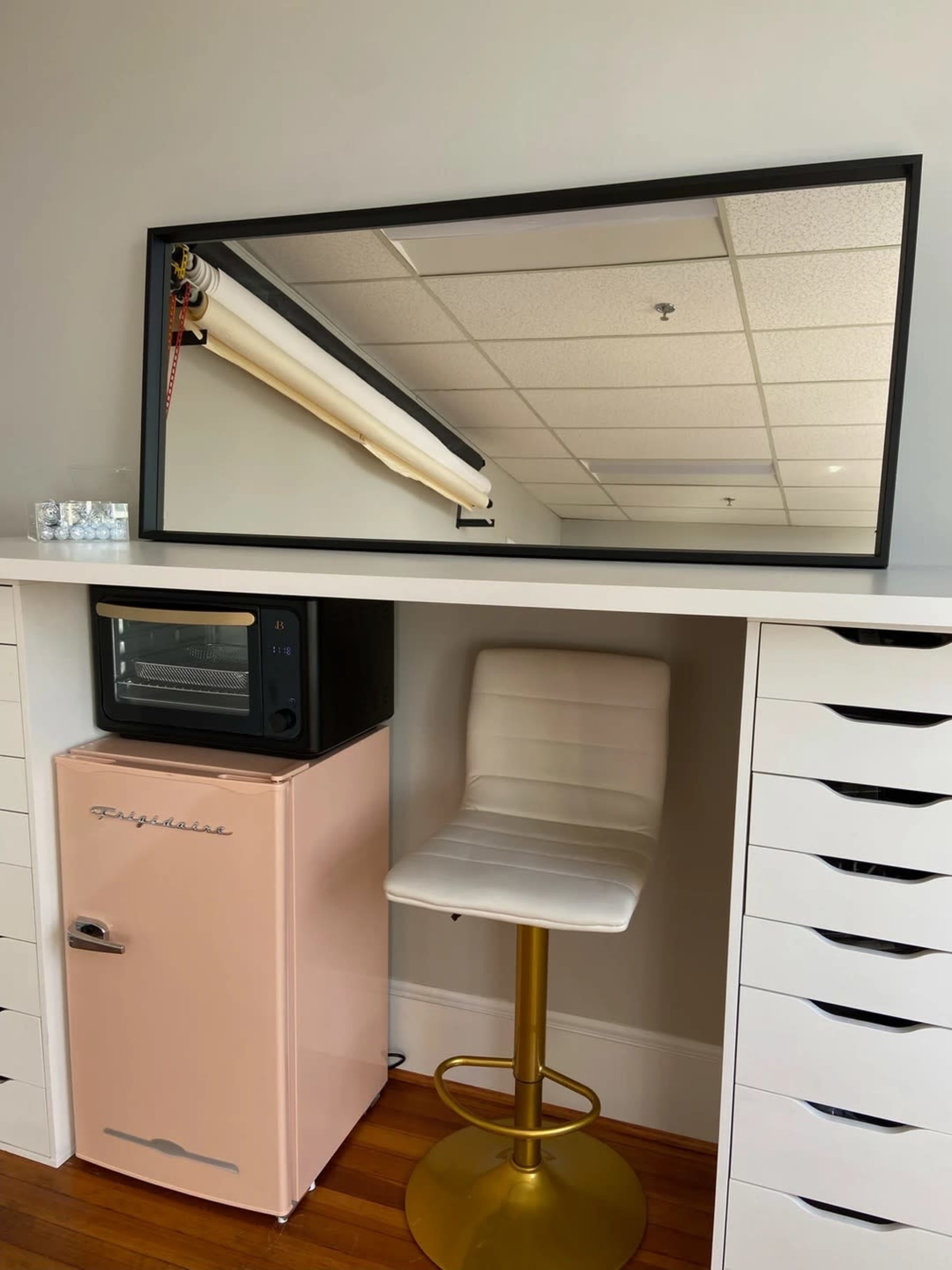 A mirror hangs above a countertop with a pink refrigerator, a black microwave, and a gold bar stool.