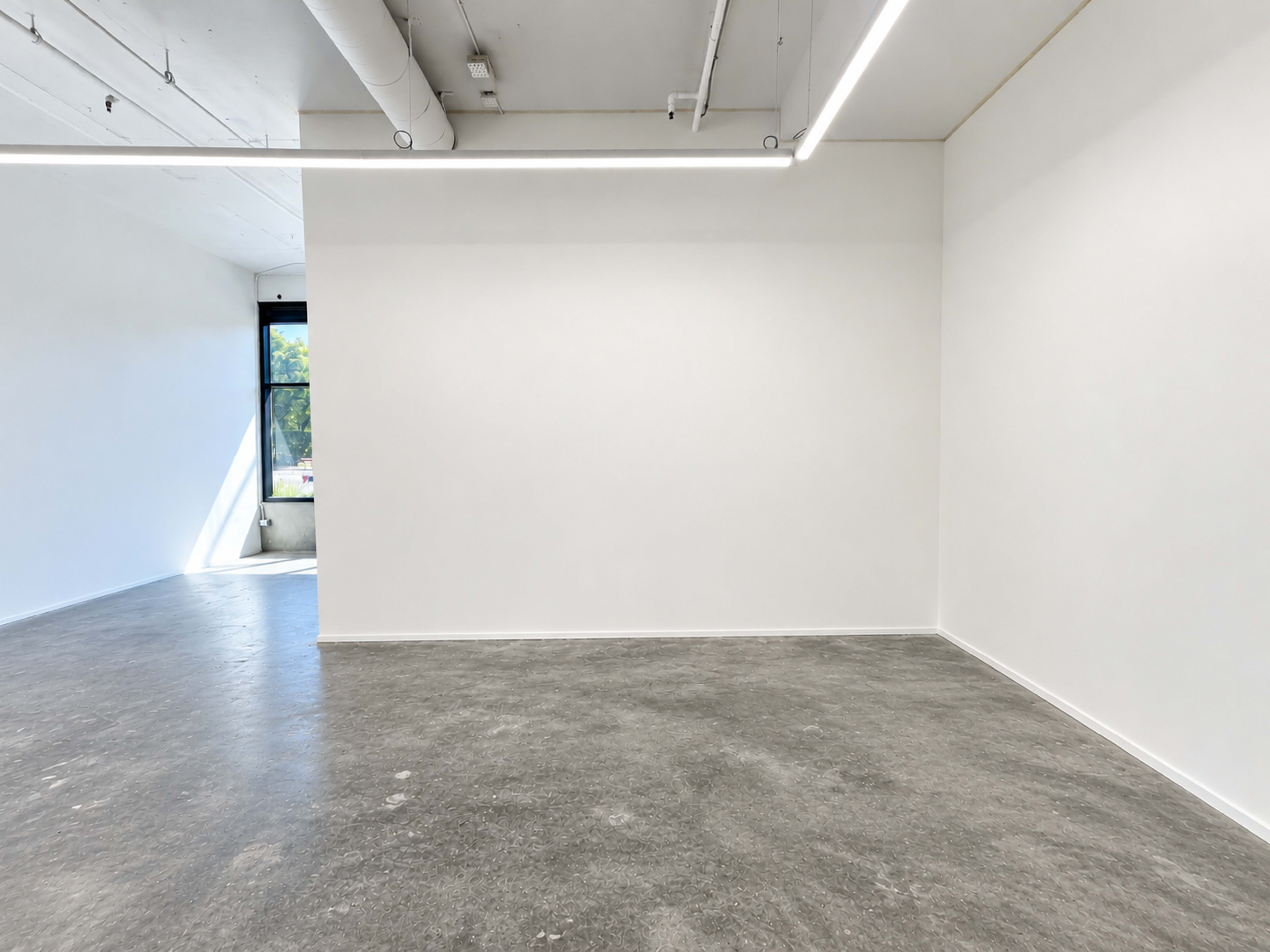 The image shows a spacious, empty room with white walls and a large window allowing natural light to enter.